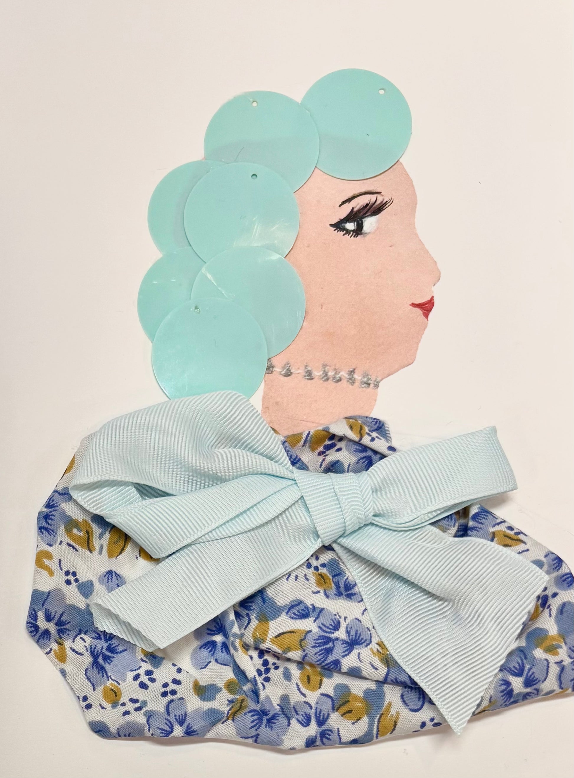 A side profile of a woman's face made with mixed materials. Her hair is created from light blue circular pieces, and she wears a silver beaded necklace. Her outfit features a floral fabric in blue, white, and yellow, topped with a large light blue ribbon bow. Her eye and lips are drawn on.