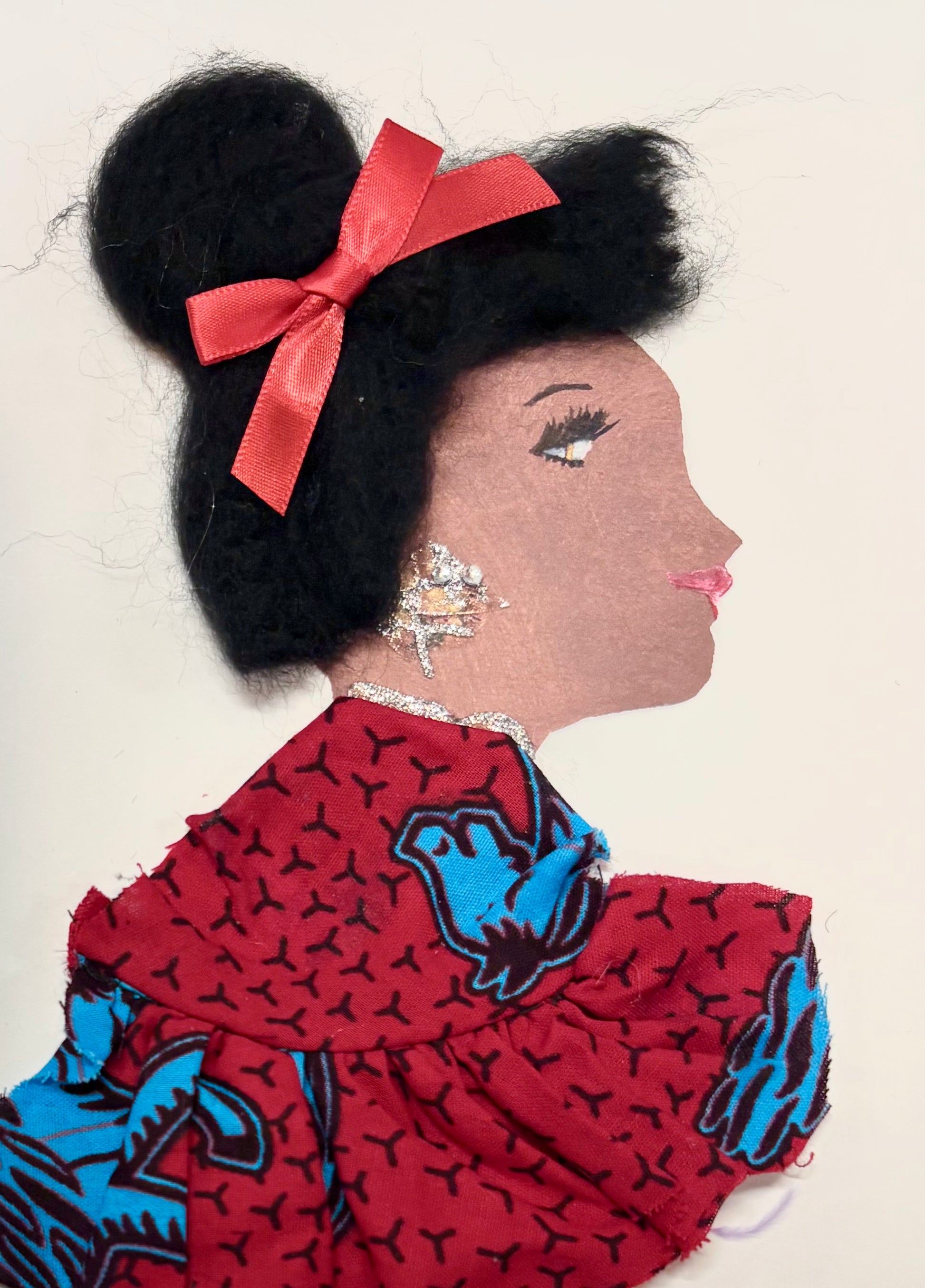 Handmade card featuring the side profile of a woman with textured black hair styled in a bun and adorned with a red ribbon. She wears a red and blue patterned fabric garment and glittery silver earrings, with a matching silver necklace. Her lips are painted pink, and she has defined eye makeup. The background is plain white.