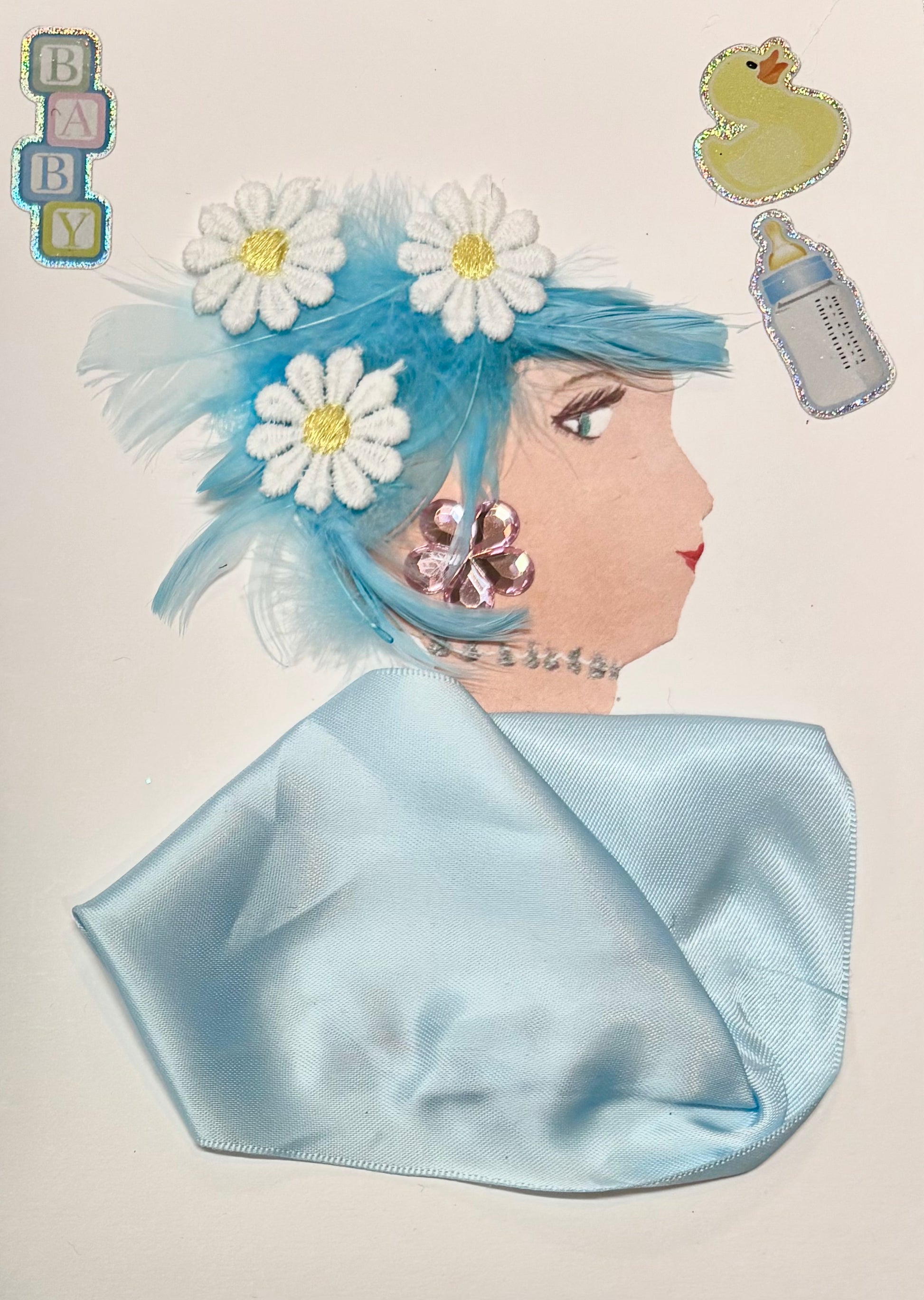 A side profile of a woman created with craft materials. She has light blue hair decorated with feathers and white daisy flowers. Her pink flower-shaped earring and silver beaded necklace add sparkle. She wears a shiny light blue fabric top. Baby-themed stickers, including the word "BABY," a rubber duck, and a bottle, appear around her head. Her eye and lips are drawn on.