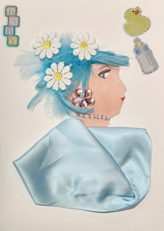 A side profile of a woman created with craft materials. She has light blue hair decorated with feathers and white daisy flowers. Her pink flower-shaped earring and silver beaded necklace add sparkle. She wears a shiny light blue fabric top. Baby-themed stickers, including the word "BABY," a rubber duck, and a bottle, appear around her head. Her eye and lips are drawn on.