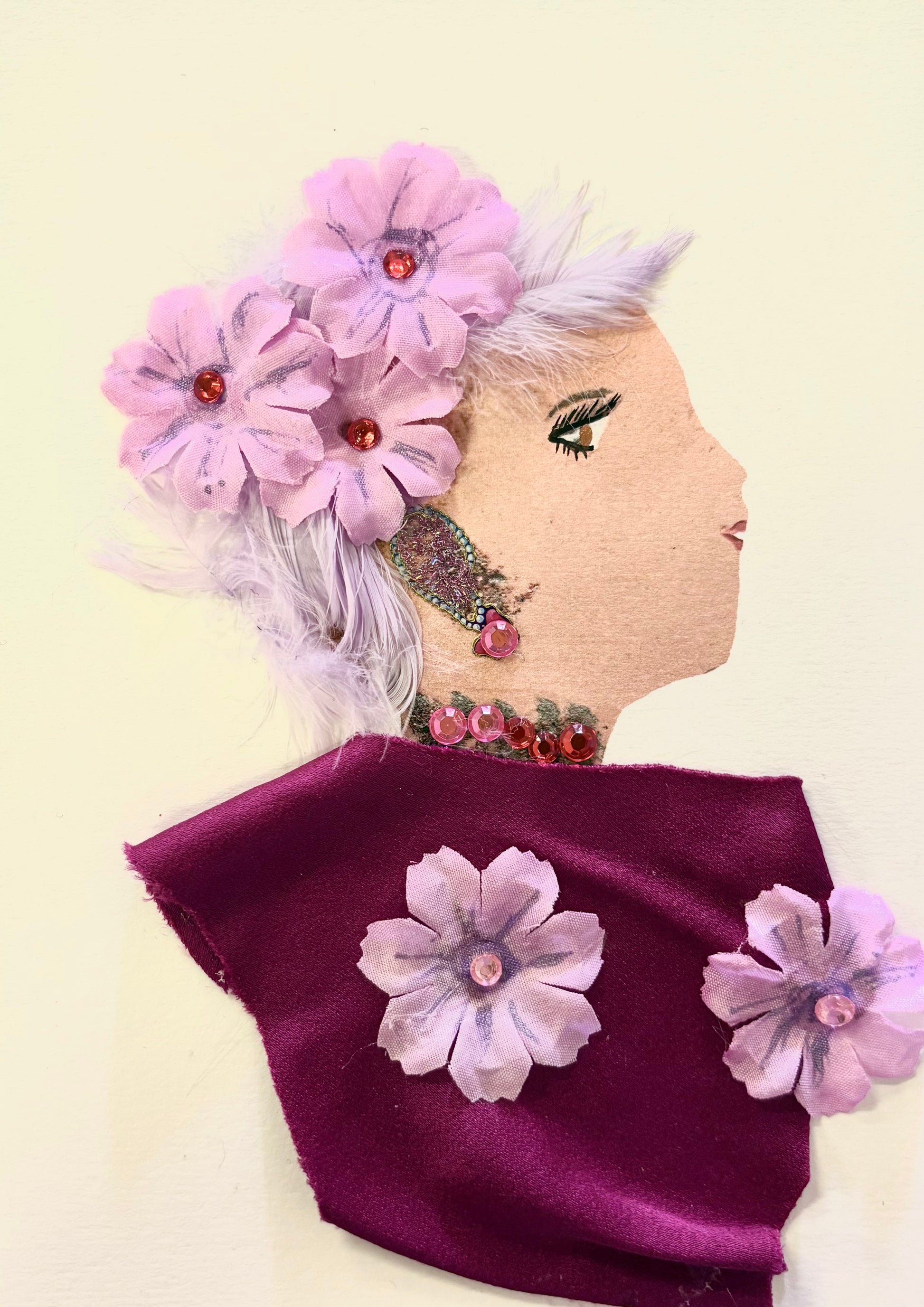 Handmade portrait of a woman in profile, crafted using mixed media. The woman has pale purple feathered hair adorned with three pink fabric flowers featuring red gem centres. She wears a deep maroon fabric garment decorated with similar fabric flowers and pink sequins as jewellery on her neck and ear. Her face is drawn on a beige textured surface, with defined eyeliner and dark lipstick.