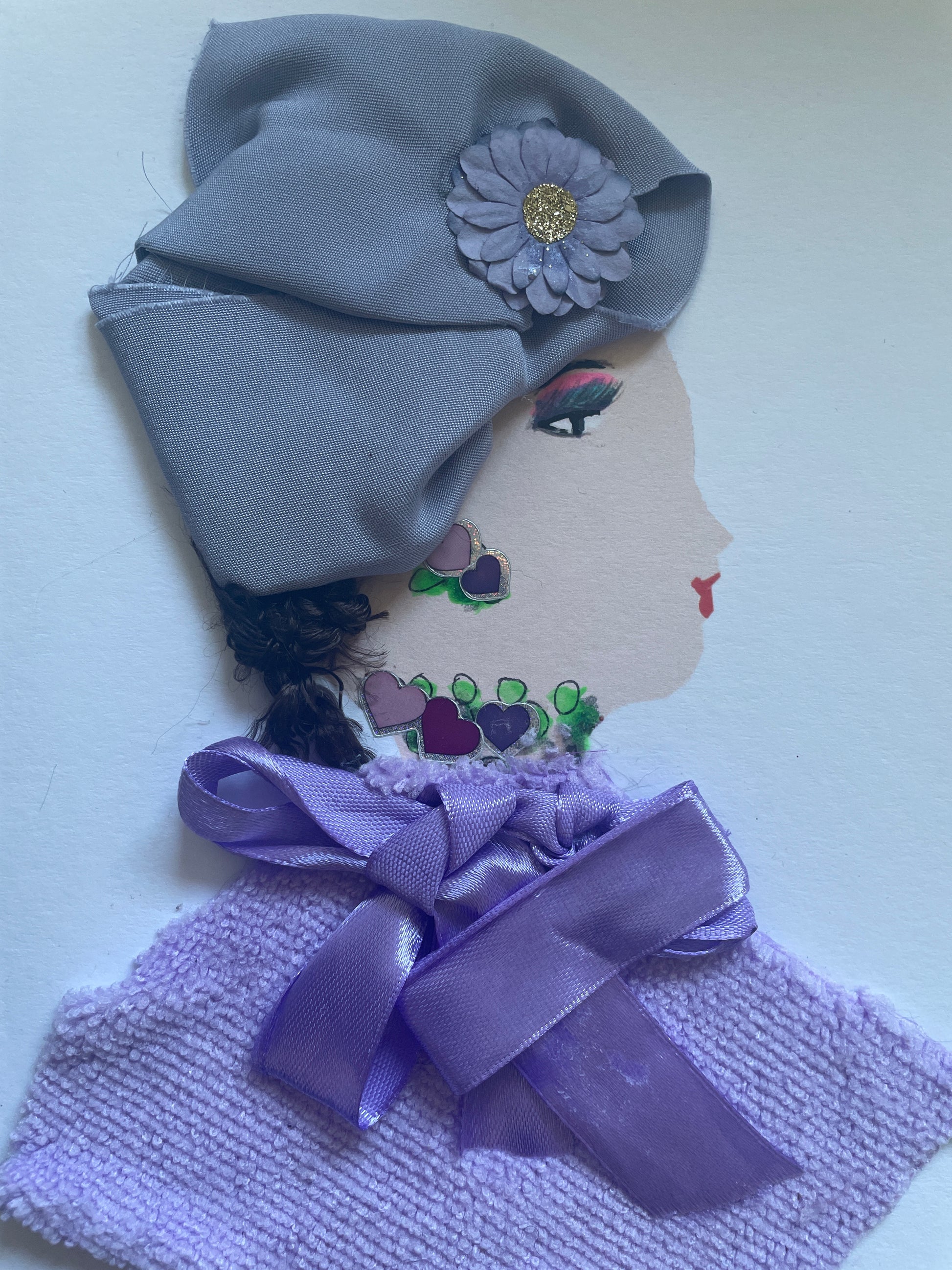 This is a handmade card of a woman named Julie Jolly. She wears a soft lavender headwrap decorated with a fabric flower that has a glittery gold centre. Her earring and necklace are painted with heart shapes in shades of purple and pink, surrounded by green beads. She is dressed in a textured lavender top with a satin bow at the collar. Her face is shown in profile, with red lips and eyeshadow in purple and green tones.