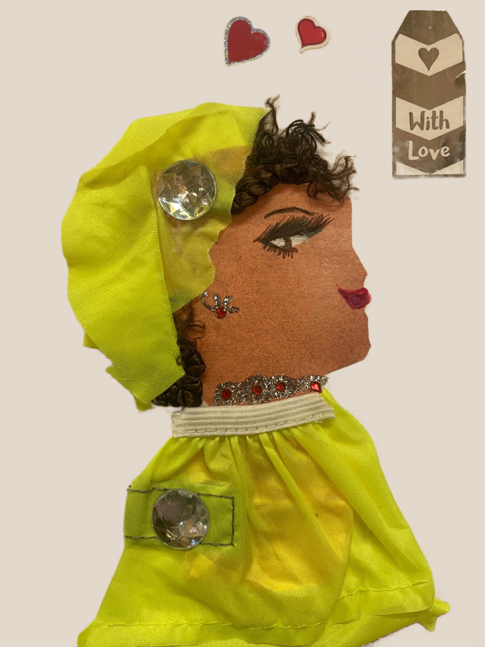 This is a handmade card of a woman named Keira Wembley. She wears a neon yellow outfit and wrap, each decorated with large clear gem buttons. She has curly dark hair, a red gem earring, and matching embellishments on her neckline. In the background are two red heart stickers and a silver tag that says "With Love."