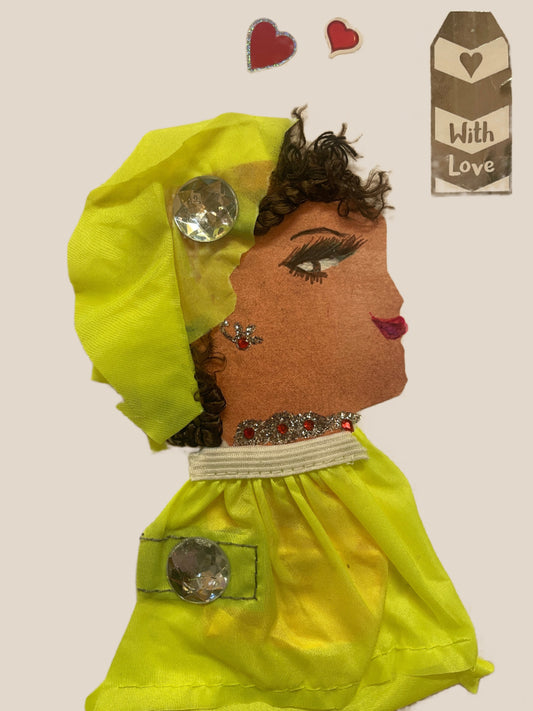 This is a handmade card of a woman named Keira Wembley. She wears a neon yellow outfit and wrap, each decorated with large clear gem buttons. She has curly dark hair, a red gem earring, and matching embellishments on her neckline. In the background are two red heart stickers and a silver tag that says "With Love."