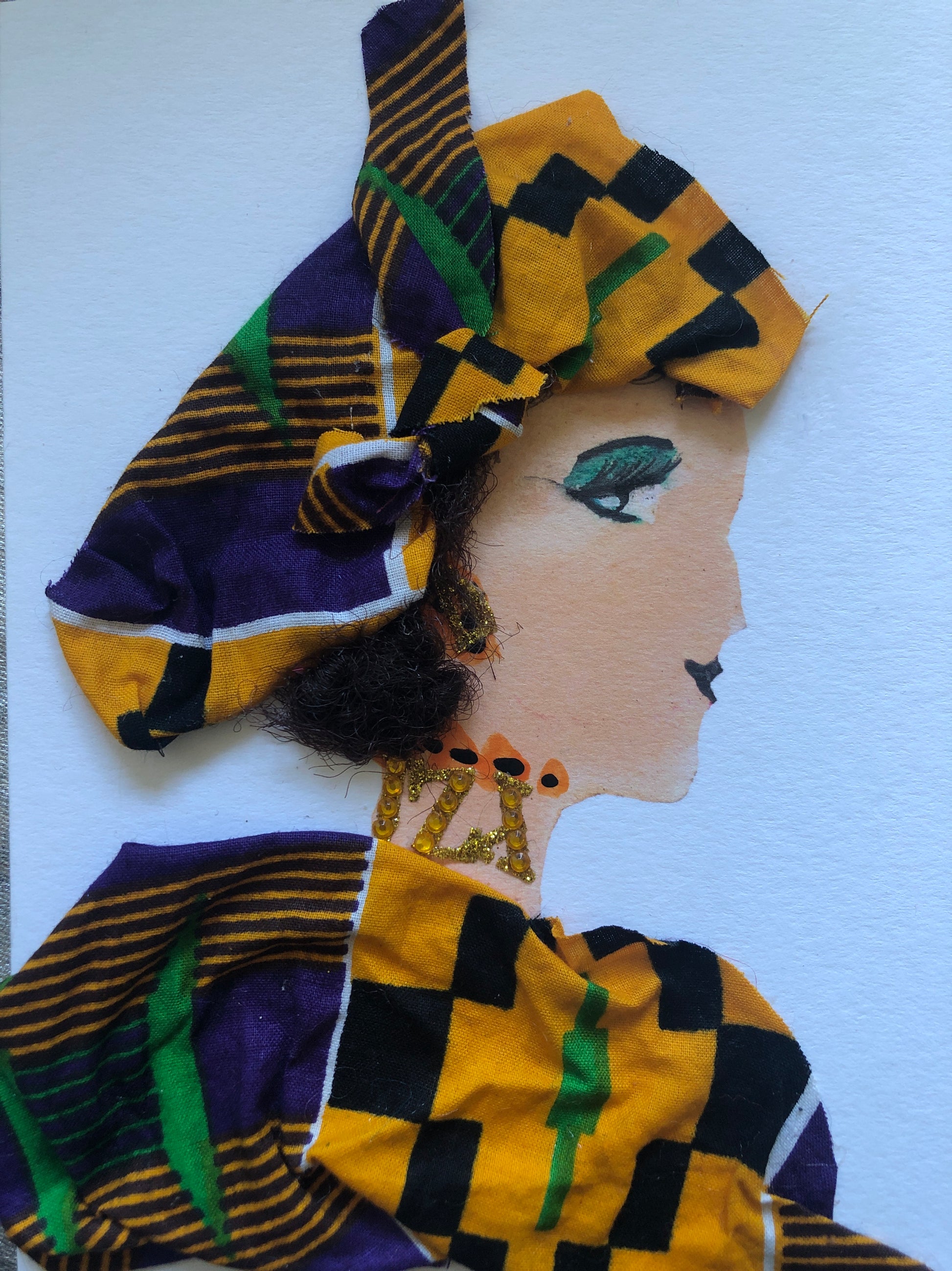 I handmade this card of a woman named Kente Wife. She’s shown in profile wearing a bold headwrap and garment made from vibrant yellow, green, black, and purple Kente-style fabric. Her eyeshadow is a striking emerald green, and her jewellery sparkles with golden glitter and orange accents. A soft textured bun completes her elegant, traditional look.