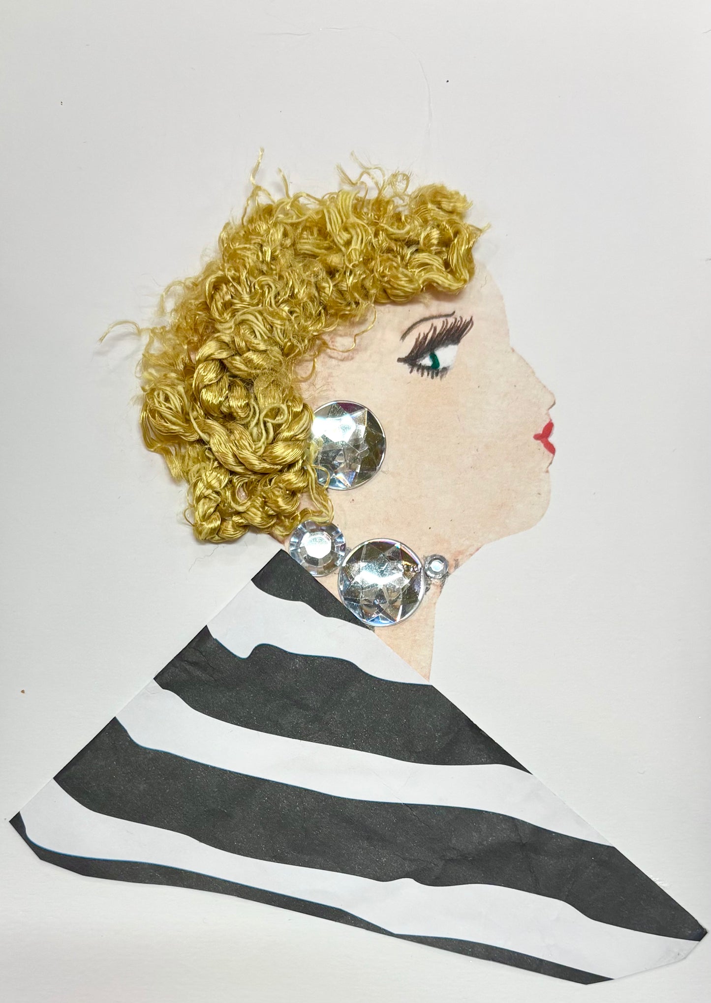 I handmade this card of a woman named Kingsbury Kira. She’s shown in profile with curly golden yarn hair, red lips, and dramatic lashes. Her jewellery sparkles with oversized clear gems along her ear and neckline. She wears a bold black-and-white striped outfit that adds to her striking, modern style.