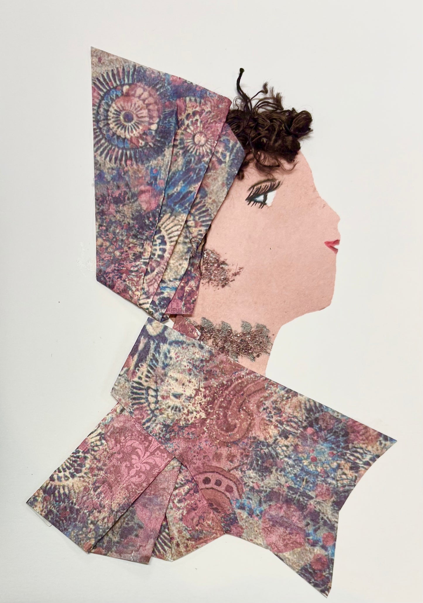 A woman wears a multicoloured pointed hat with her brown hair peaking out at the top. She has a matching blouse with the same multicoloured pattern featuring pinks, blues, and yellows. She wears sparkling jewellery.