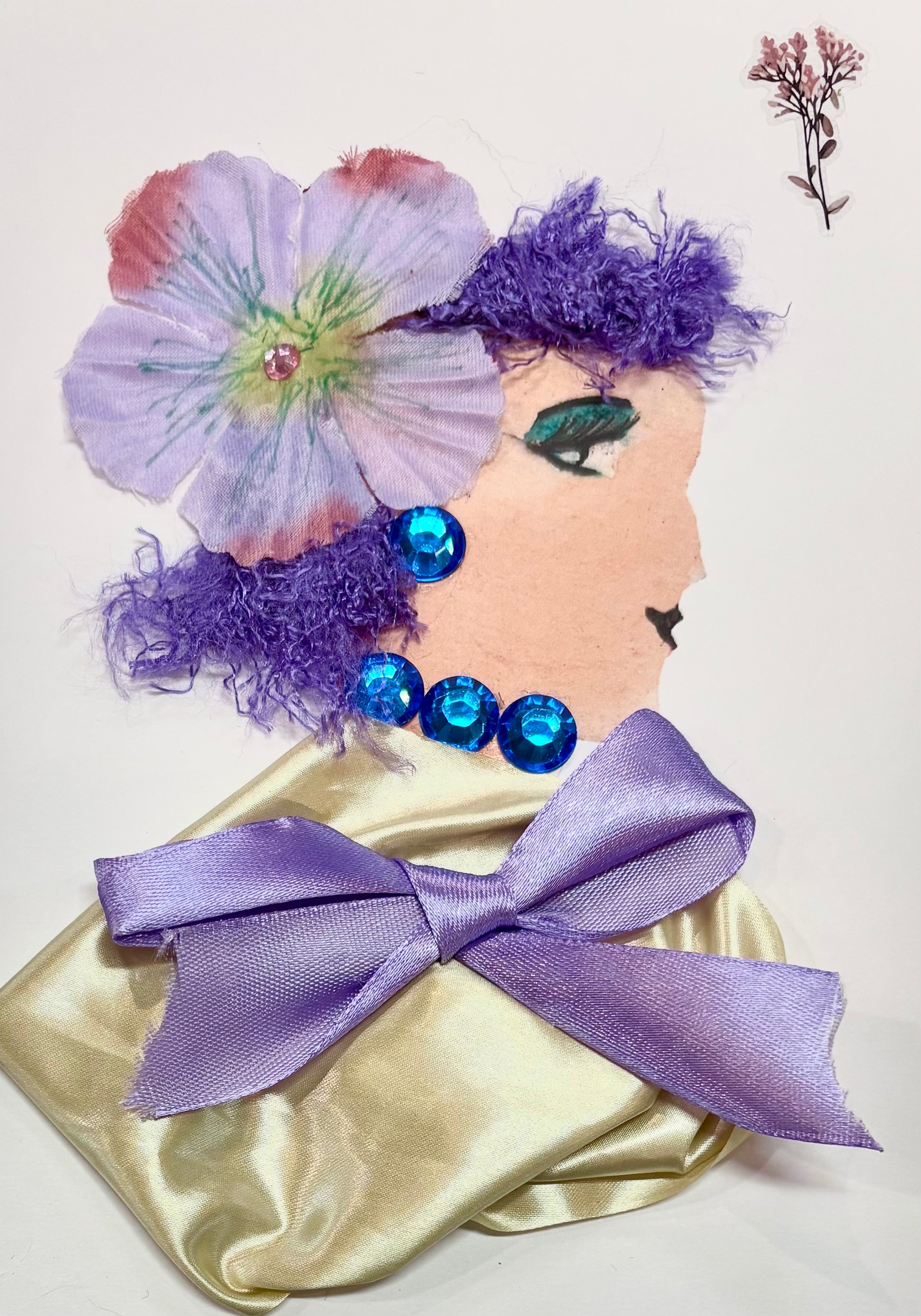 A woman has dark purple hair with a light purple flower as an accessory. She wears a gold blouse with a light purple bow placed in the center. She has sapphire blue gem earrings and a necklace. A dainty flower sits in the top right corner of the card.