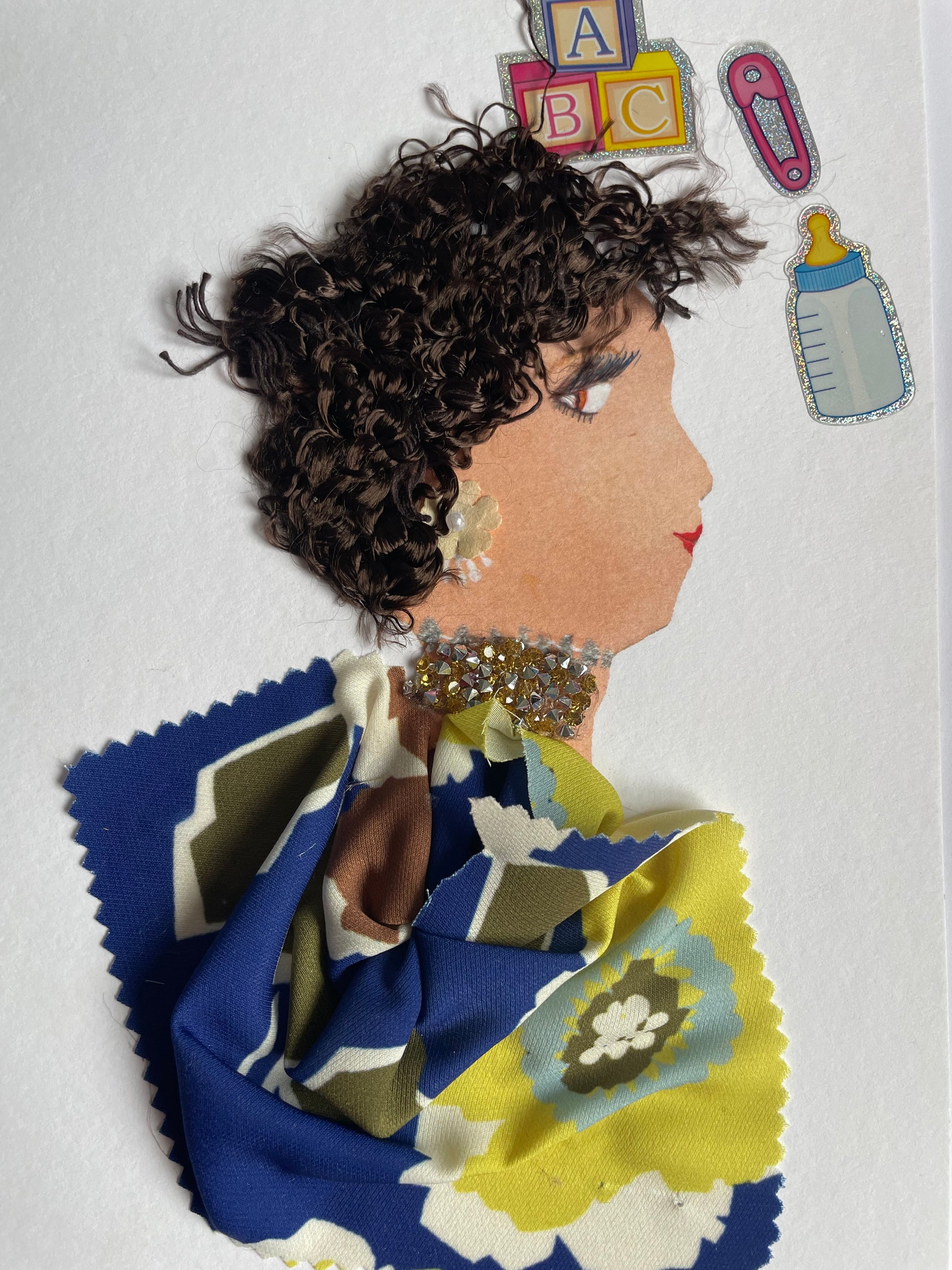 I handmade this card of a woman named Lambeth Lucy. She’s shown in profile with curly brown yarn hair, red lips, and soft makeup. Her gold-toned earring and glittering neckline add sparkle to her look. She wears a vibrant floral-patterned fabric in blue, green, yellow, and cream. Around her are baby-themed stickers, ABC blocks, a bottle, and a dummy clip, suggesting a nurturing, playful spirit.
