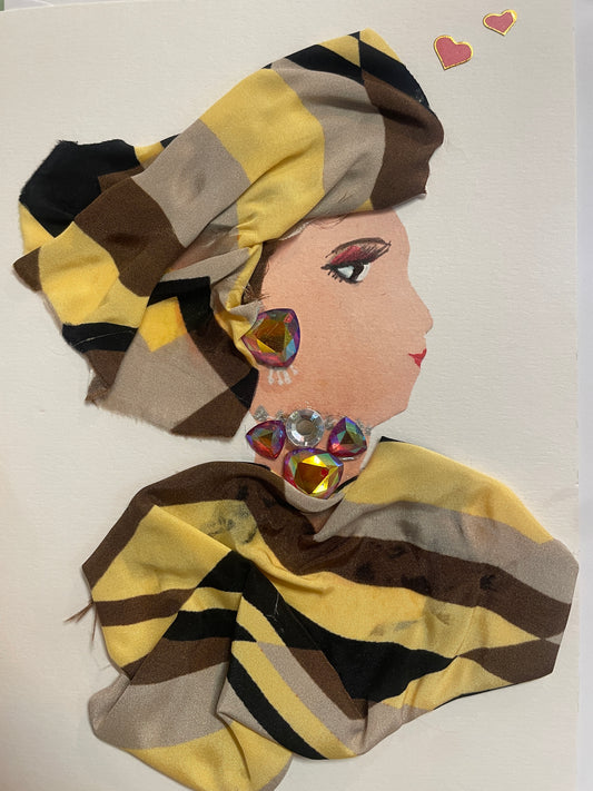 This is a handmade card of a woman named Leyla Ludgate. She wears a striped turban and matching fabric wrap in tones of yellow, brown, and black, with bold gem earrings and neckline decorations. Two heart stickers are in the background.