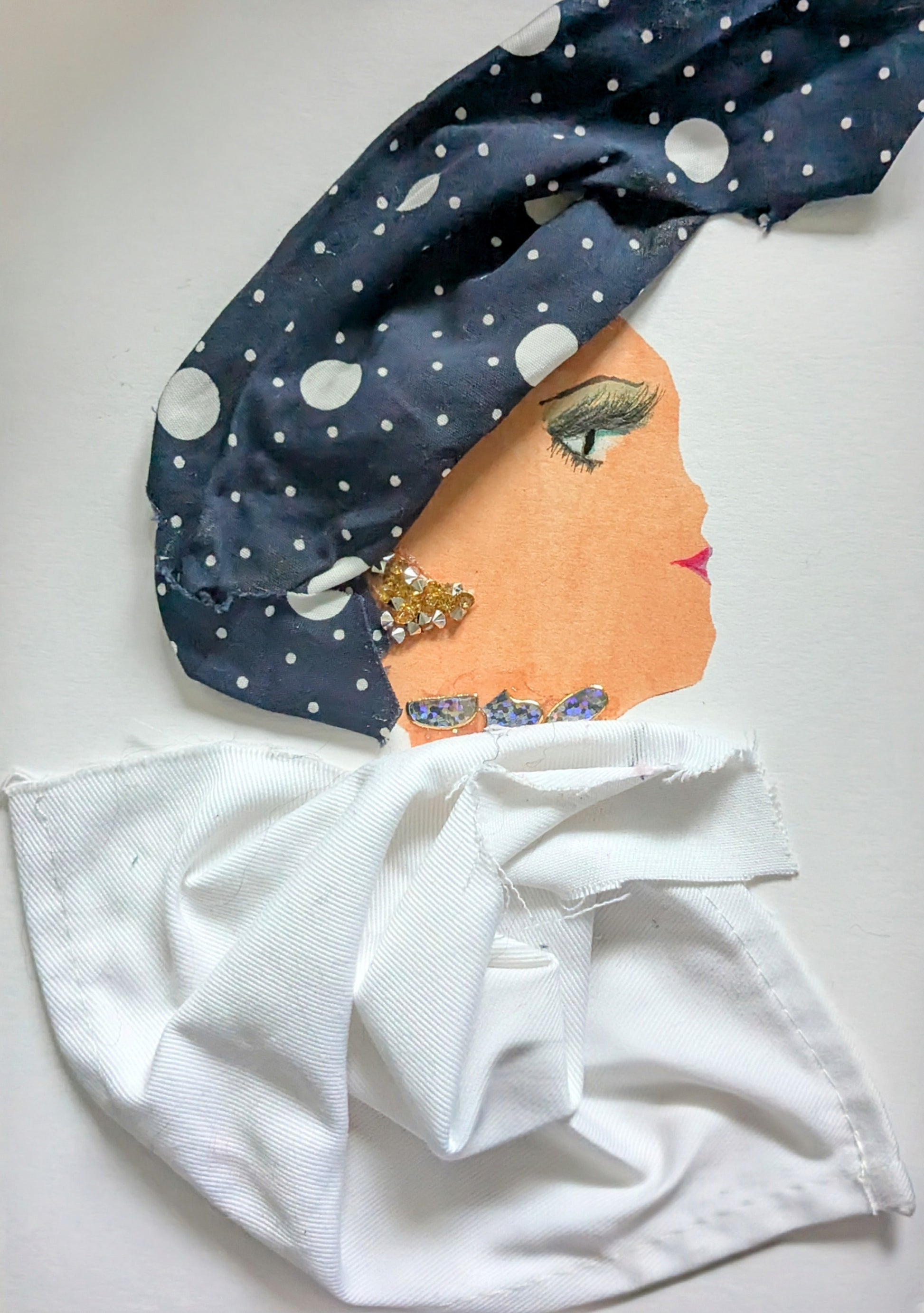 I handmade this card of a woman named London Lisa. She’s shown in profile with bright pink lips, bold eyeliner, and a confident gaze. Her headwrap is made from navy blue fabric with scattered white polka dots, and she wears sparkly gold and crystal-like earrings. A shimmering detail wraps around her neck, and her crisp white outfit is fashioned from folded cloth.