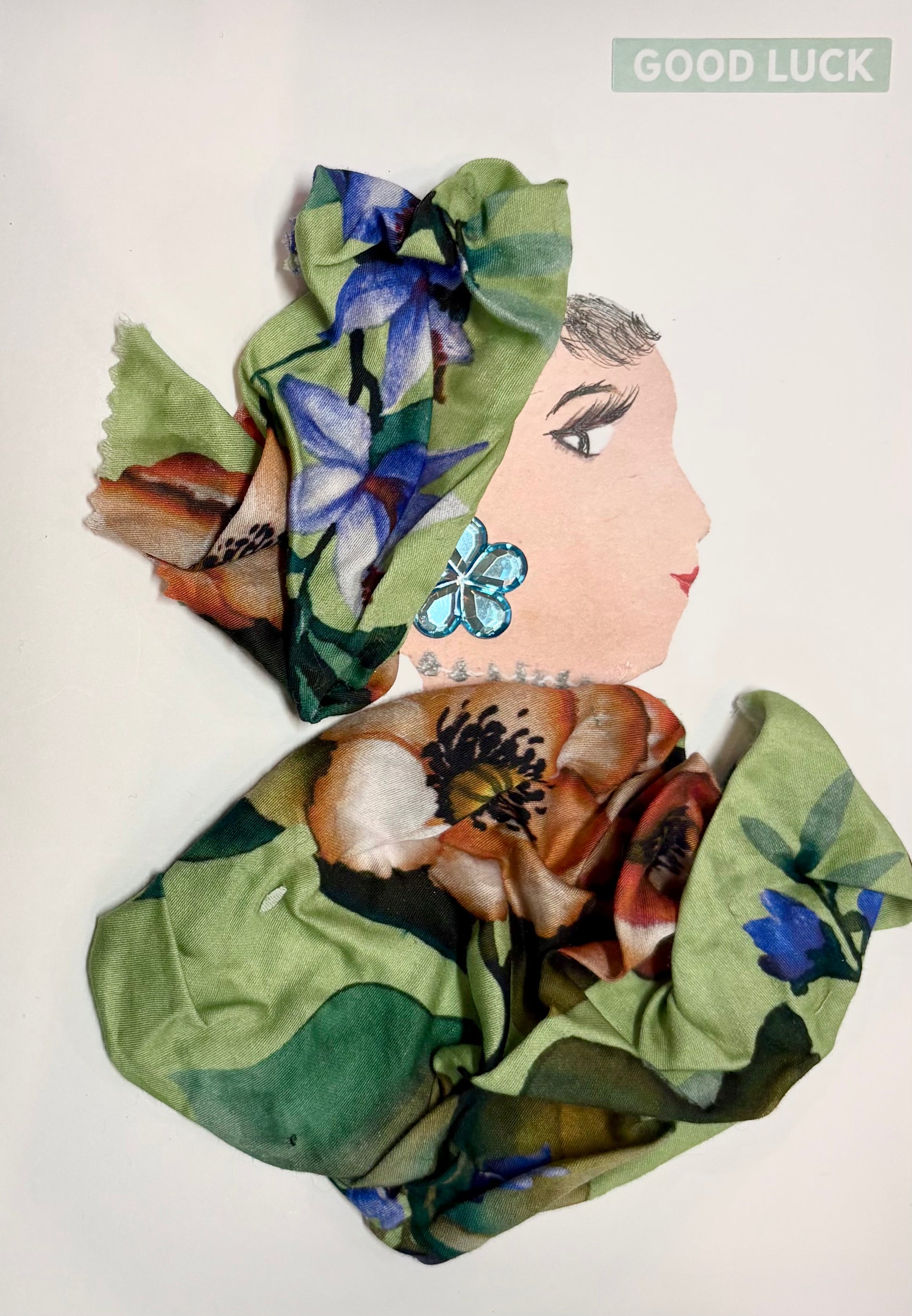 Here we have Lucky Lucy, cloaked in a garden of blossoms. Her outfit and headwrap are crafted from lush green fabric adorned with vibrant floral prints—orange, purple, and blue petals bloom all around her. A glimmering blue flower earring and a delicate jeweled necklace add sparkle to her look. In the corner, a soft “GOOD LUCK” message floats