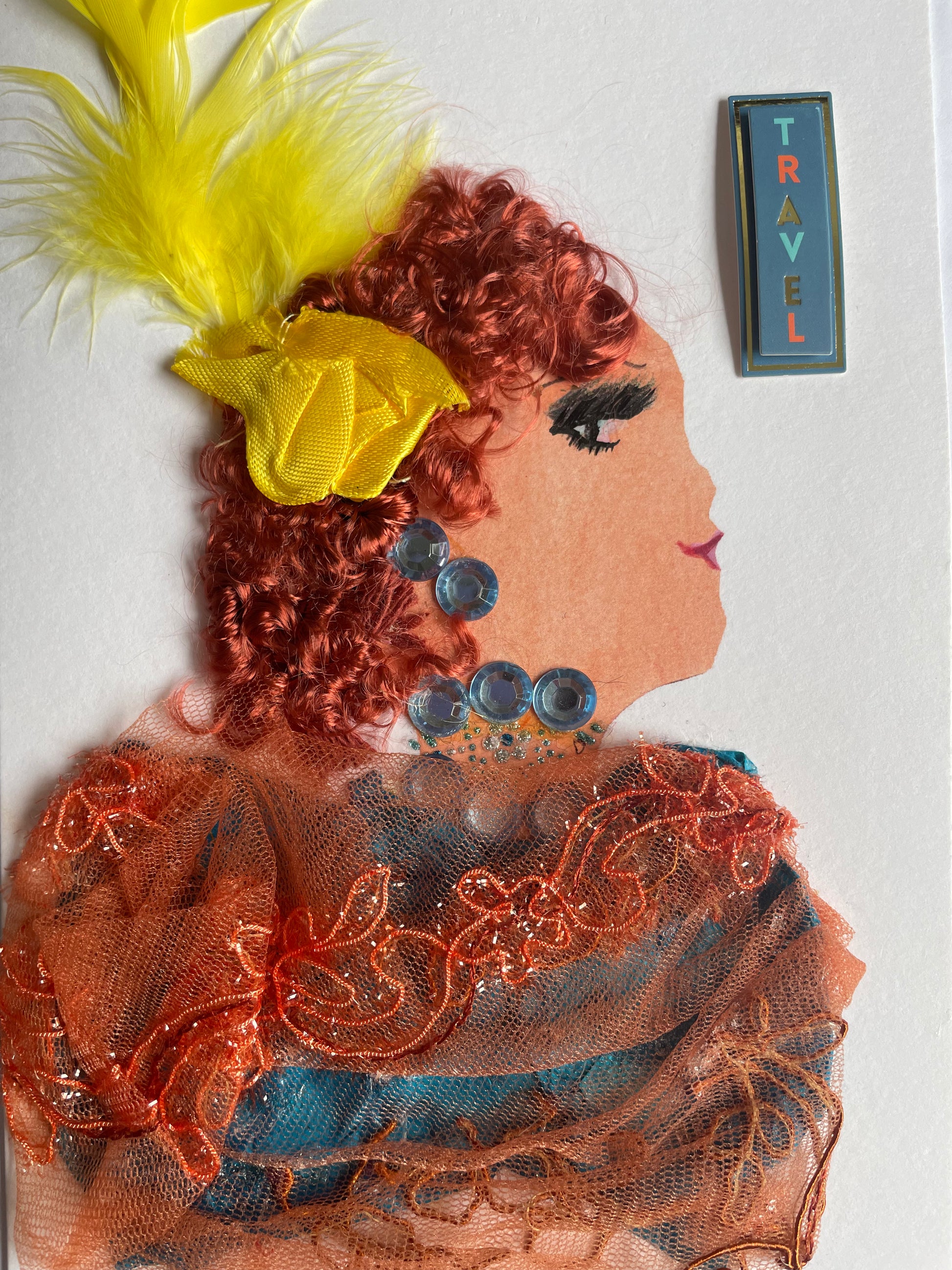 I handmade this card of a woman named Maritime Madison. She’s shown in profile with curly auburn hair, dramatic eye makeup, and a soft smile. She wears sparkling blue gems on her ear and neckline, a bright yellow bow with feathers in her hair, and a layered outfit made from orange embroidered mesh over teal fabric. A sticker beside her reads “TRAVEL” in colourful block letters.
