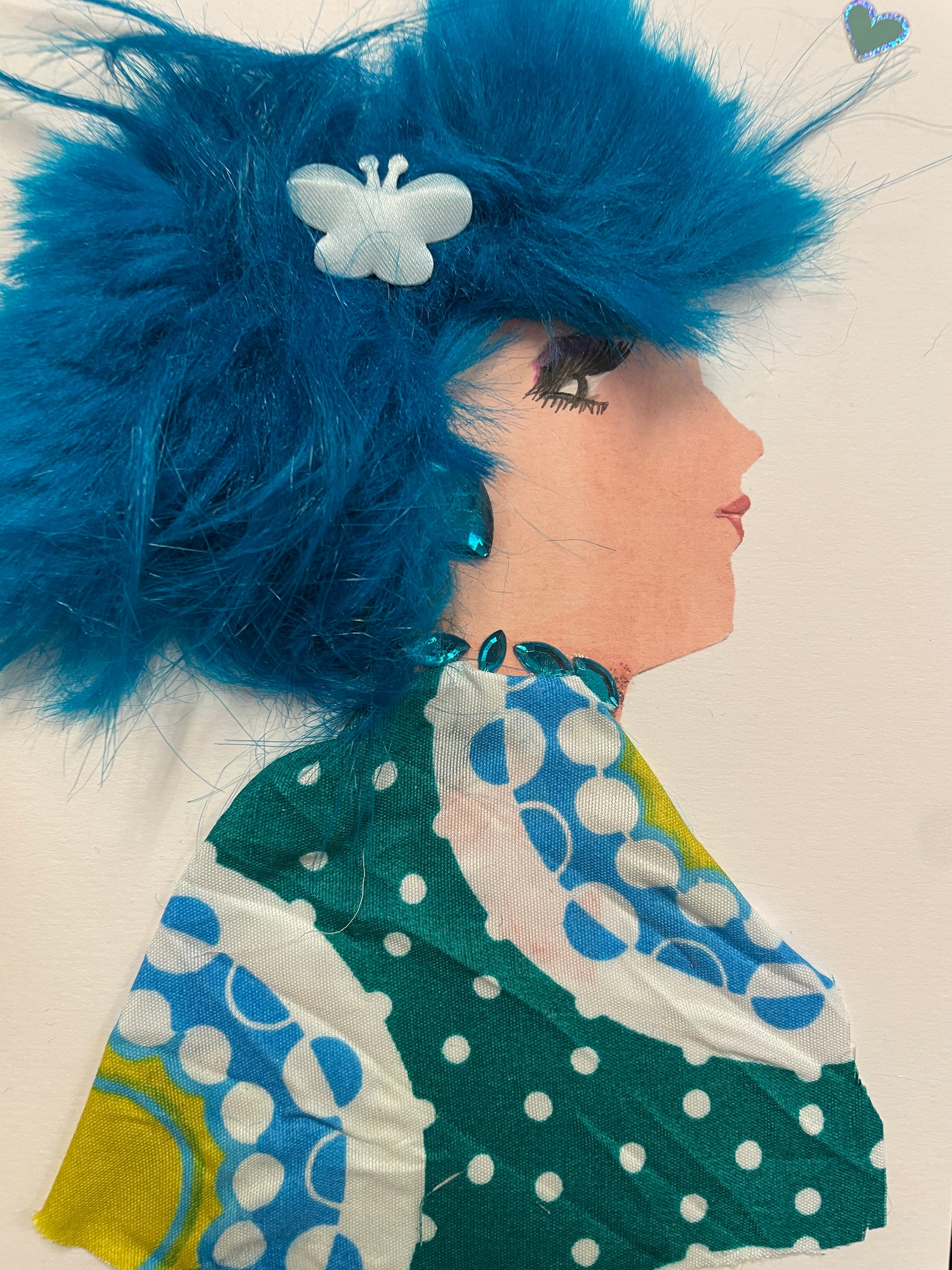 Handmade card of a woman in profile with fluffy teal-blue hair, decorated with a white butterfly clip. She wears a colorful green, blue, and white polka-dot dress and blue gemstone accents at the neckline. A small holographic heart sticker floats in the top right corner