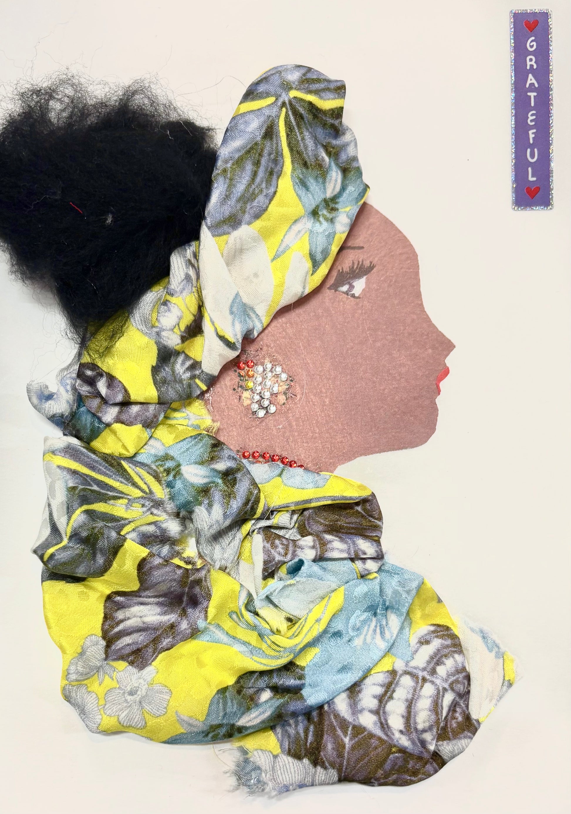 I handmade this card of a woman named Mellow Yellow. She wears a large, flowing headwrap and matching outfit made from soft fabric with yellow, blue, and grey floral patterns. Her hair is styled in a full, textured bun. She has a sparkling earring made of small red and silver gems and a red beaded necklace. A small label beside her reads “GRATEFUL.”
