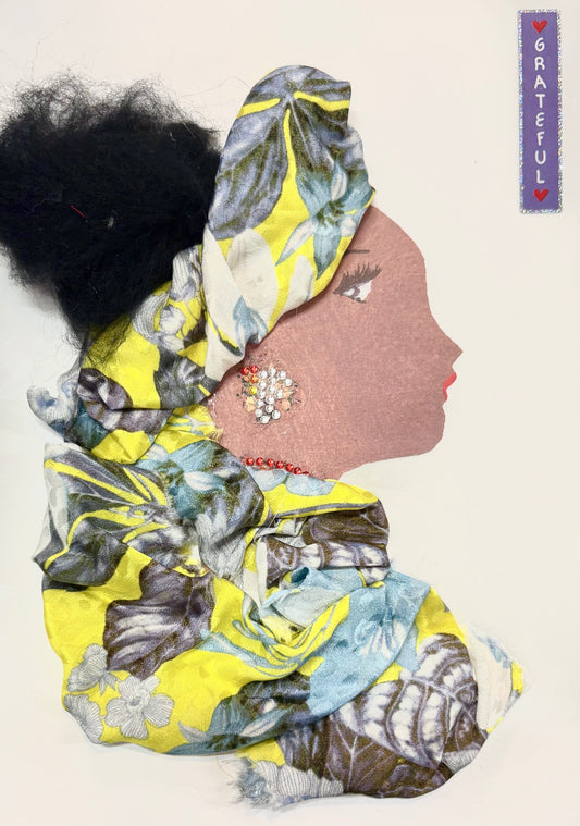 I handmade this card of a woman named Mellow Yellow. She wears a large, flowing headwrap and matching outfit made from soft fabric with yellow, blue, and grey floral patterns. Her hair is styled in a full, textured bun. She has a sparkling earring made of small red and silver gems and a red beaded necklace. A small label beside her reads “GRATEFUL.”
