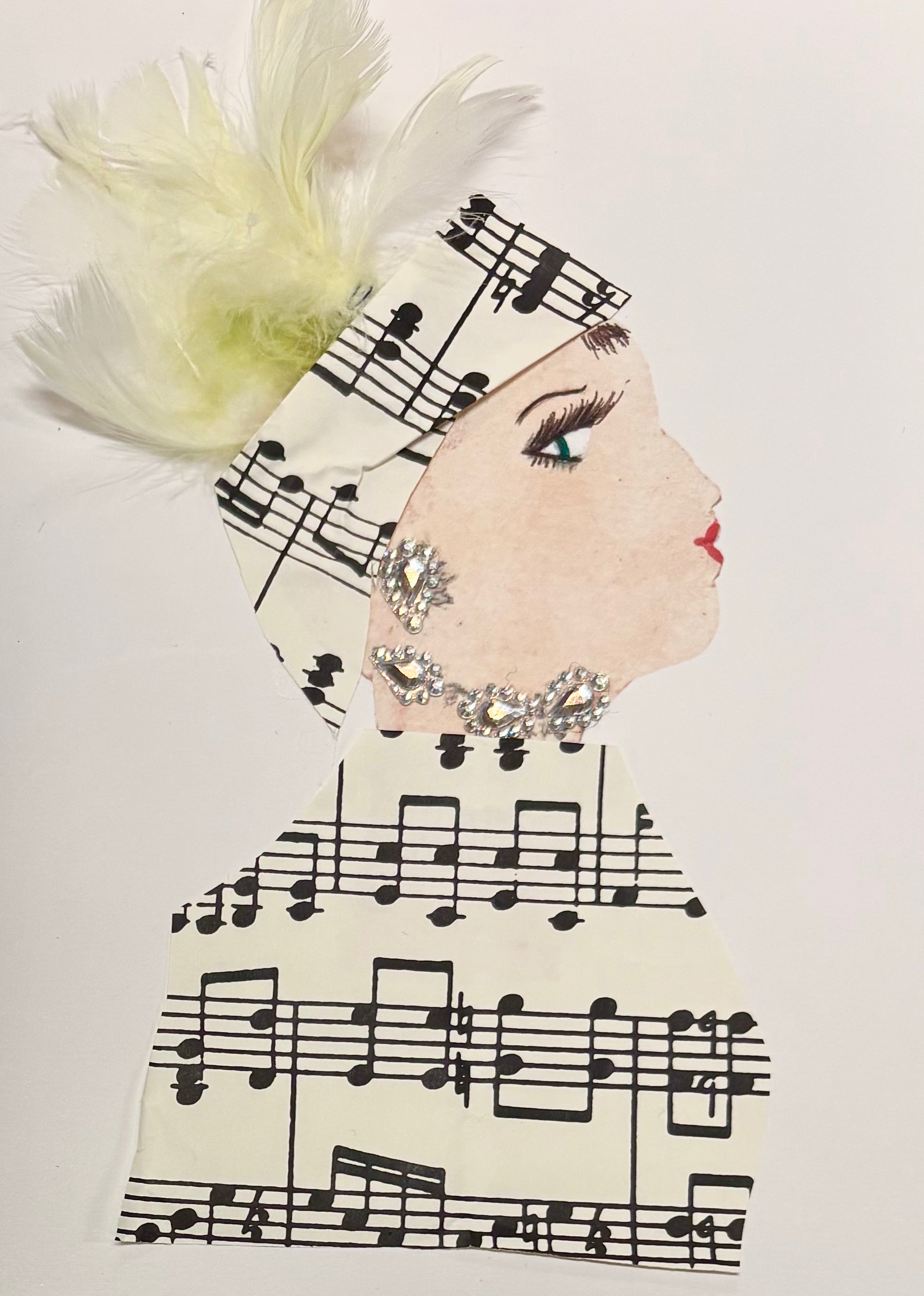 I handmade this card of a woman named Melody Music. She’s shown in profile with defined lashes, red lips, and a serene expression. Her headwrap and outfit are made from sheet music paper, and pale yellow feathers plume from the top of her wrap. She’s adorned with glittering silver rhinestones as earrings and at her neck, echoing her elegant, melodic style.