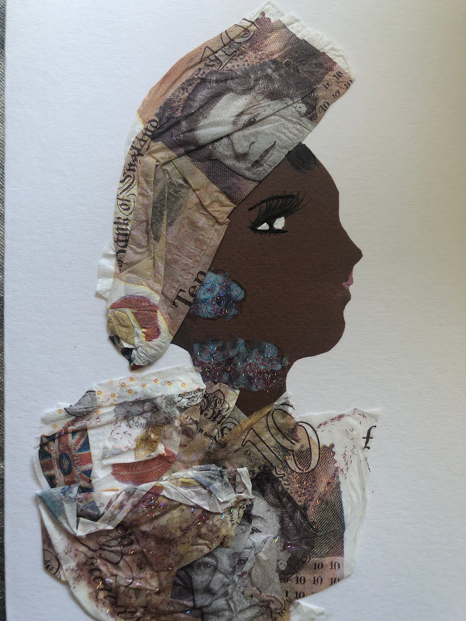 This is a handmade card of a woman named Monica Pounds. Her face is a deep brown tone. Her hair wrap and clothing are made from textured, crumpled paper resembling British pound notes. She wears glittery blue earrings and a matching necklace.