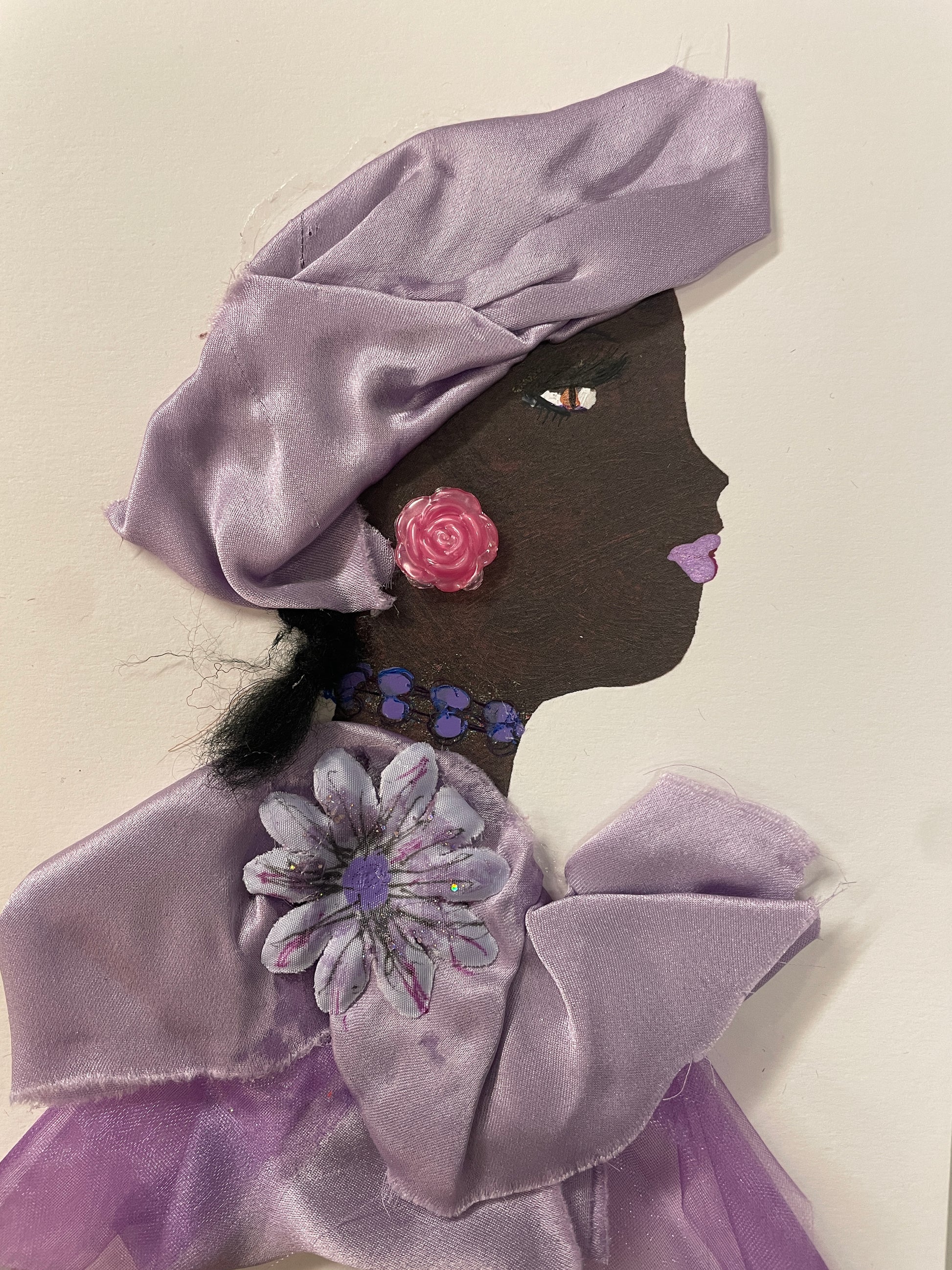 This is a handmade card of a woman named Nia Blossom. She wears a shiny purple outfit with pink and white flowers, a cluster of pearls at the neck, and has short curly hair.