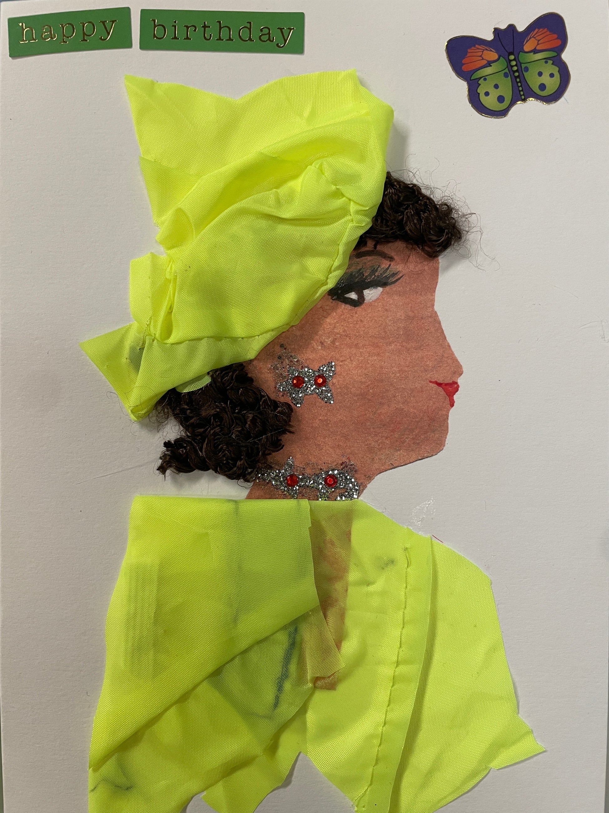 This is a handmade card of a woman named Nylssa Newbury wearing a bright yellow head wrap and outfit with silver and red embellishments, and a butterfly sticker  in the top right corner. In the top left, you can see a sticker saying "happy birthday"