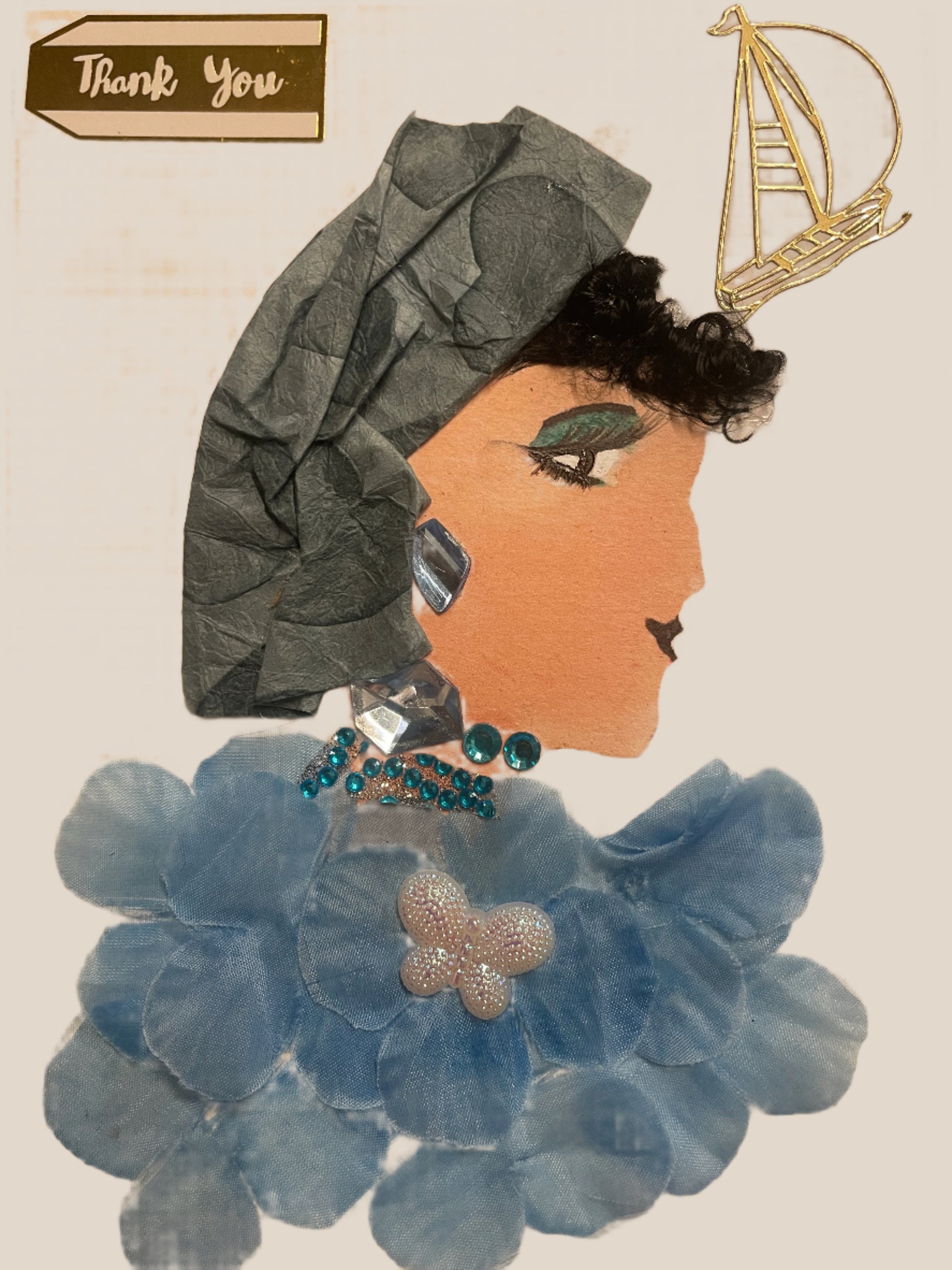 This is a handmade card of a woman named Opal Olympia. She wears a crinkled blue head wrap, curly hair, and a blouse made of layered pale blue petals. A white butterfly sits in the centre, with a “Thank You” and gold sailboat sticker above.