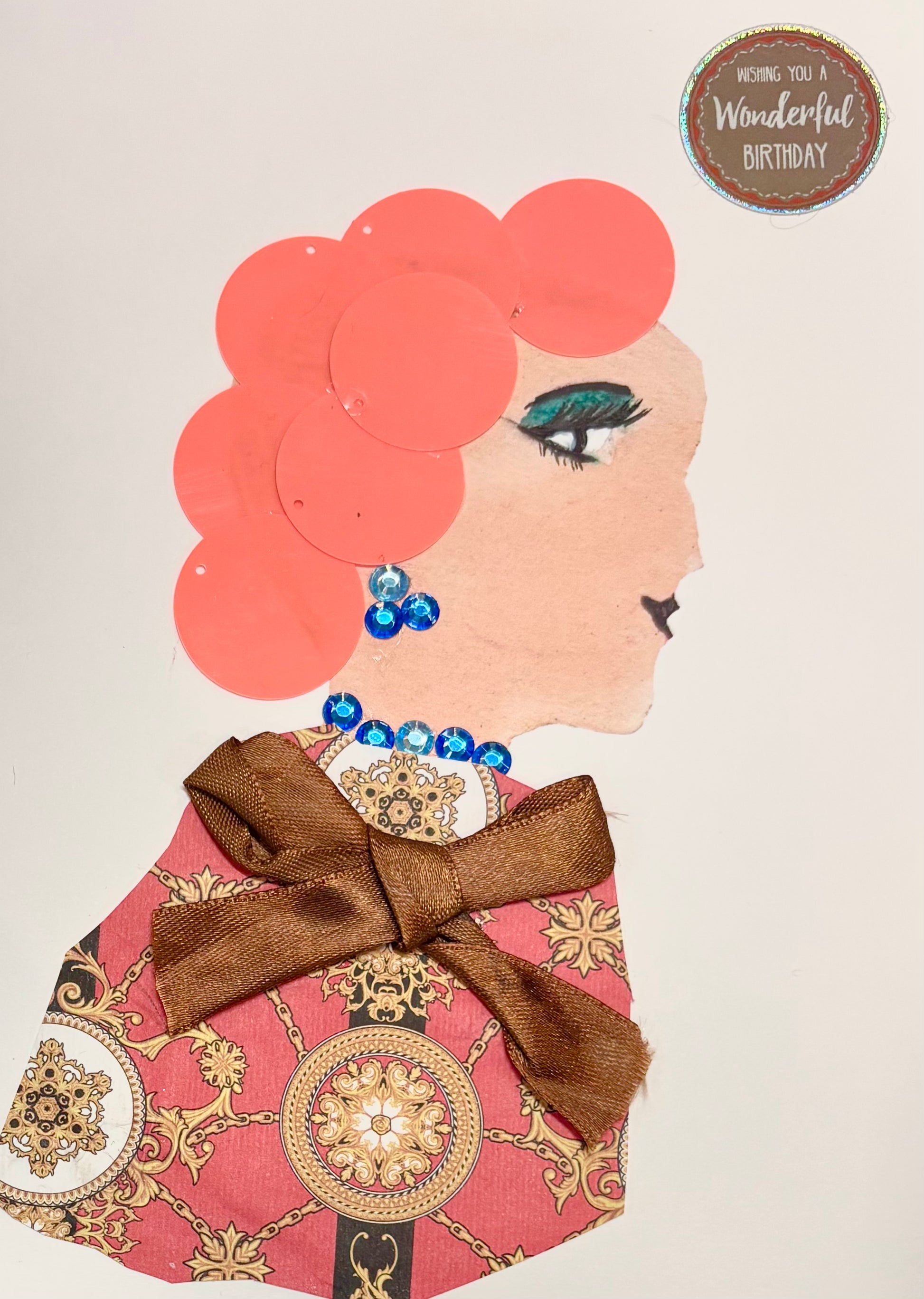A woman has subtle orange hair and a multicoloured blouse with coral, gold, and black. The blouse also features a chocolate brown bow. She wears blue gem earrings and a matching necklace. A sticker in the top right corner states, "Wishing you a wonderful birthday."