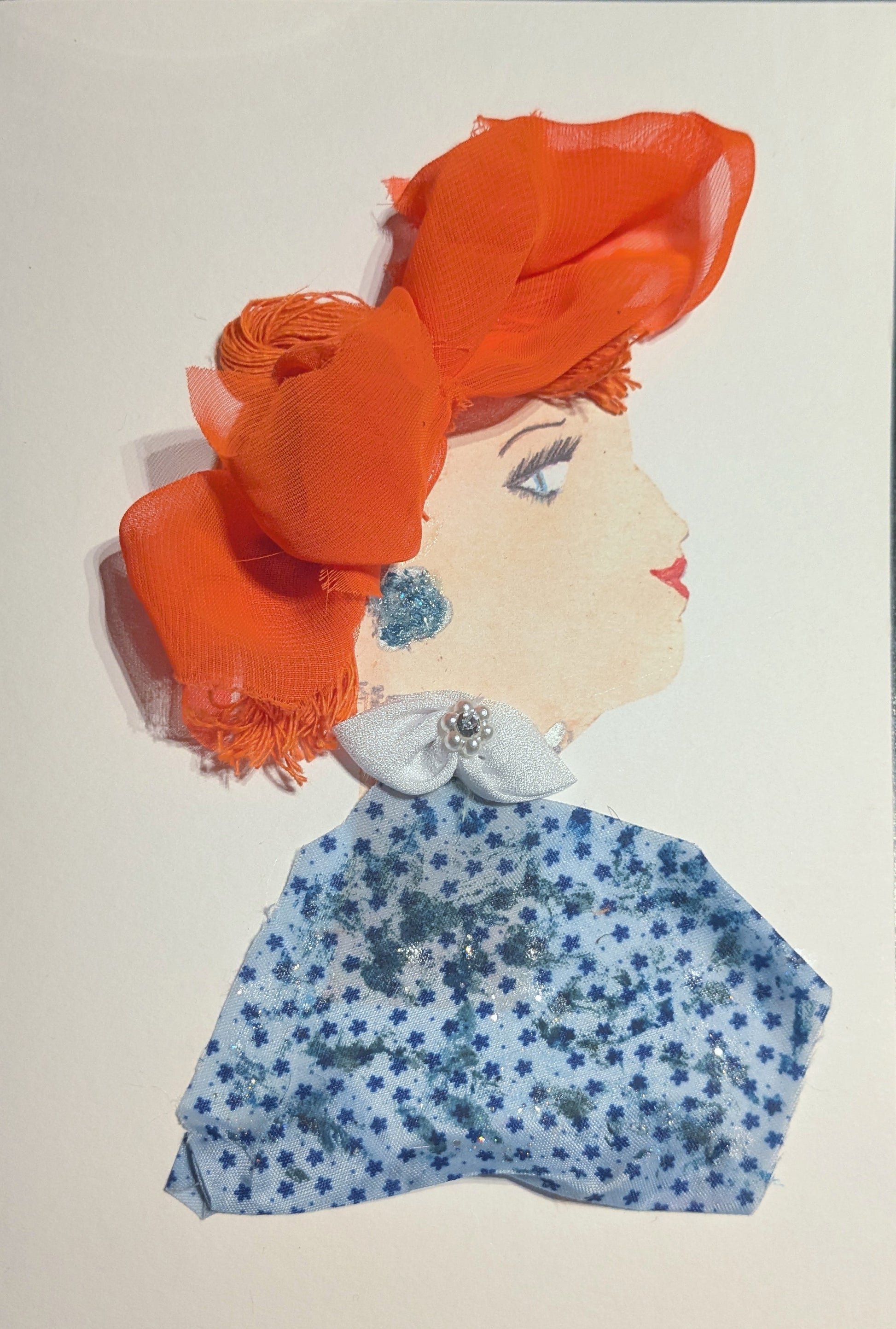 A side profile of a woman made with art materials. She has bright orange fabric styled as hair, with soft folds and texture. She wears a sparkly blue earring and a white bow with a pearl-like center at her collar. Her top is made from blue floral fabric with tiny flowers and glitter accents. Her eye and lips are drawn on.