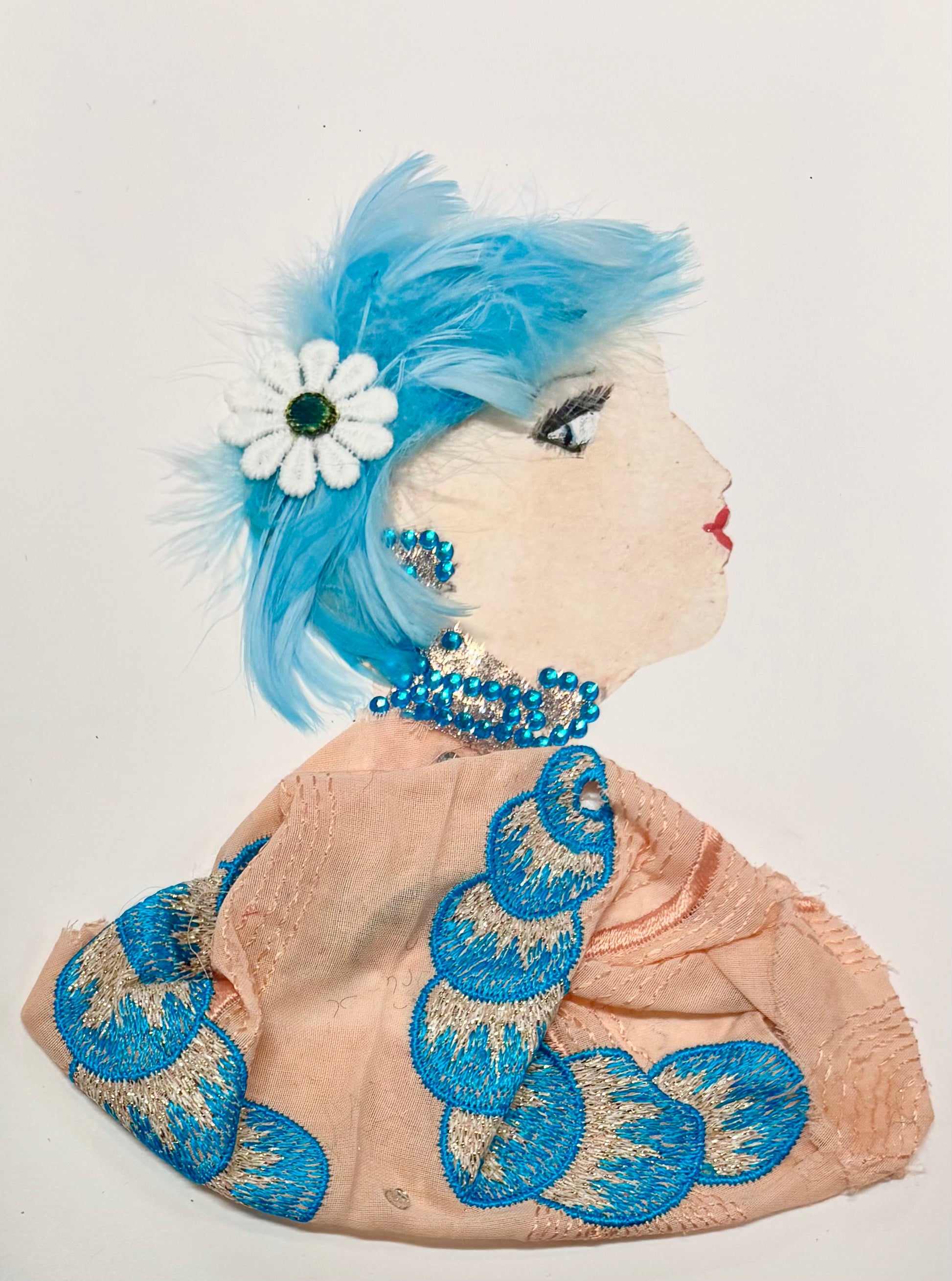This is a handmade card of a woman named Peach Penelope. She has bright blue hair made from soft feathers and yarn, decorated with a white daisy. She wears sparkling blue and silver jewellery on her ear and neck. Her outfit is made from peach-coloured fabric with bold blue and silver embroidery in a scallop pattern. Her face is shown in profile with carefully painted features and red lips.