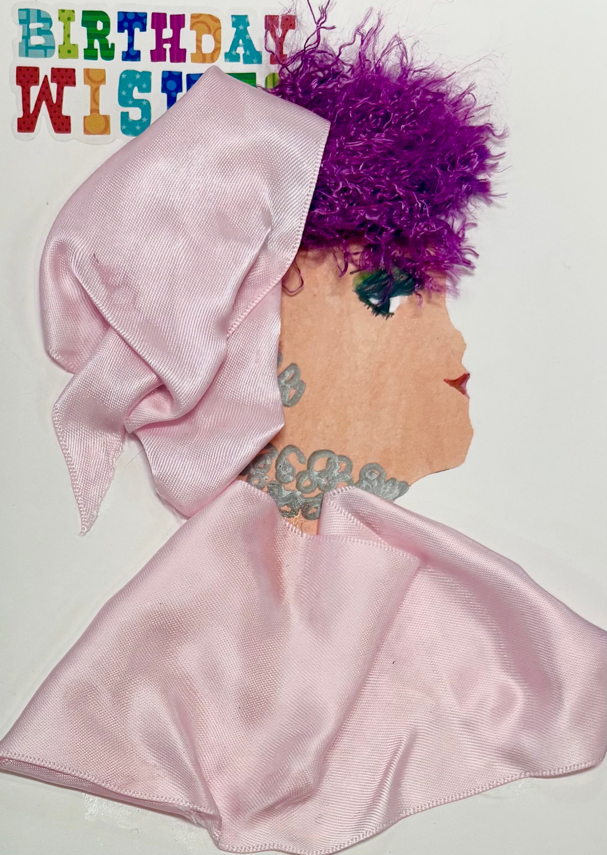 This is a handmade card of a woman named **Pink Penny**. Her bold, textured magenta curls are paired with a lustrous soft pink headwrap and matching flowing garment made from satin ribbon. Around her neck is an intricate silver-grey floral design, and the card is cheerfully accented with the words "BIRTHDAY WISHES" in multicolored lettering.