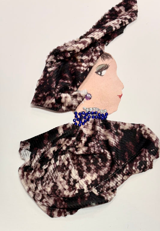 This is a handmade card of a woman named Quinn Queensbury. She wears a soft, textured fabric in maroon and cream, wrapped into a voluminous turban and matching outfit. A single purple gem sits on her ear, while her necklace is made of deep blue and silver beads, clustered closely around her neck for a regal touch.