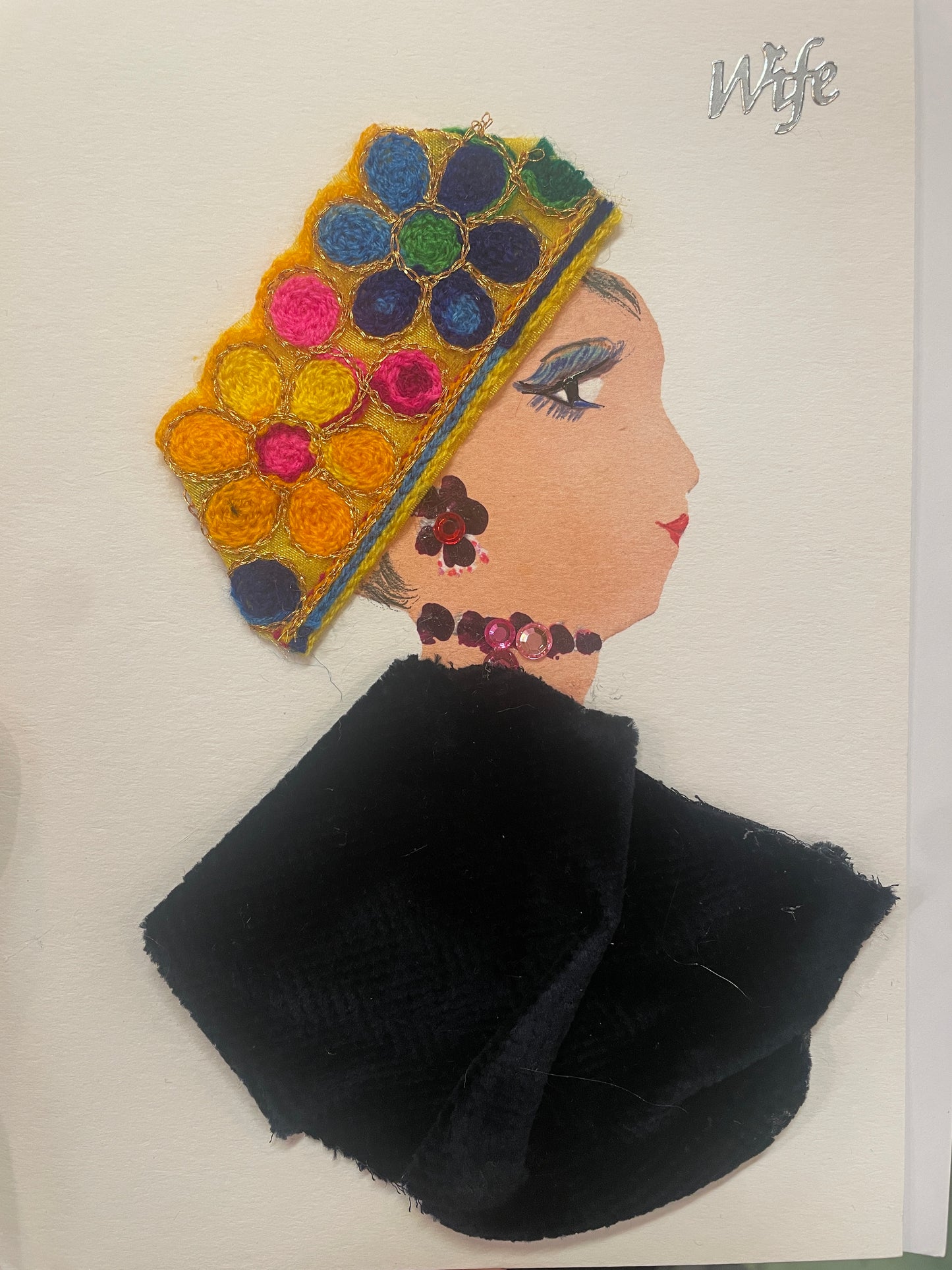 This is a handmade card of a woman named Regent Renee. She wears a textured yellow hat adorned with multicoloured yarn flowers, a black velvet cloak, and pink beaded accessories. The word “Wife” is written in silver in the top right corner.