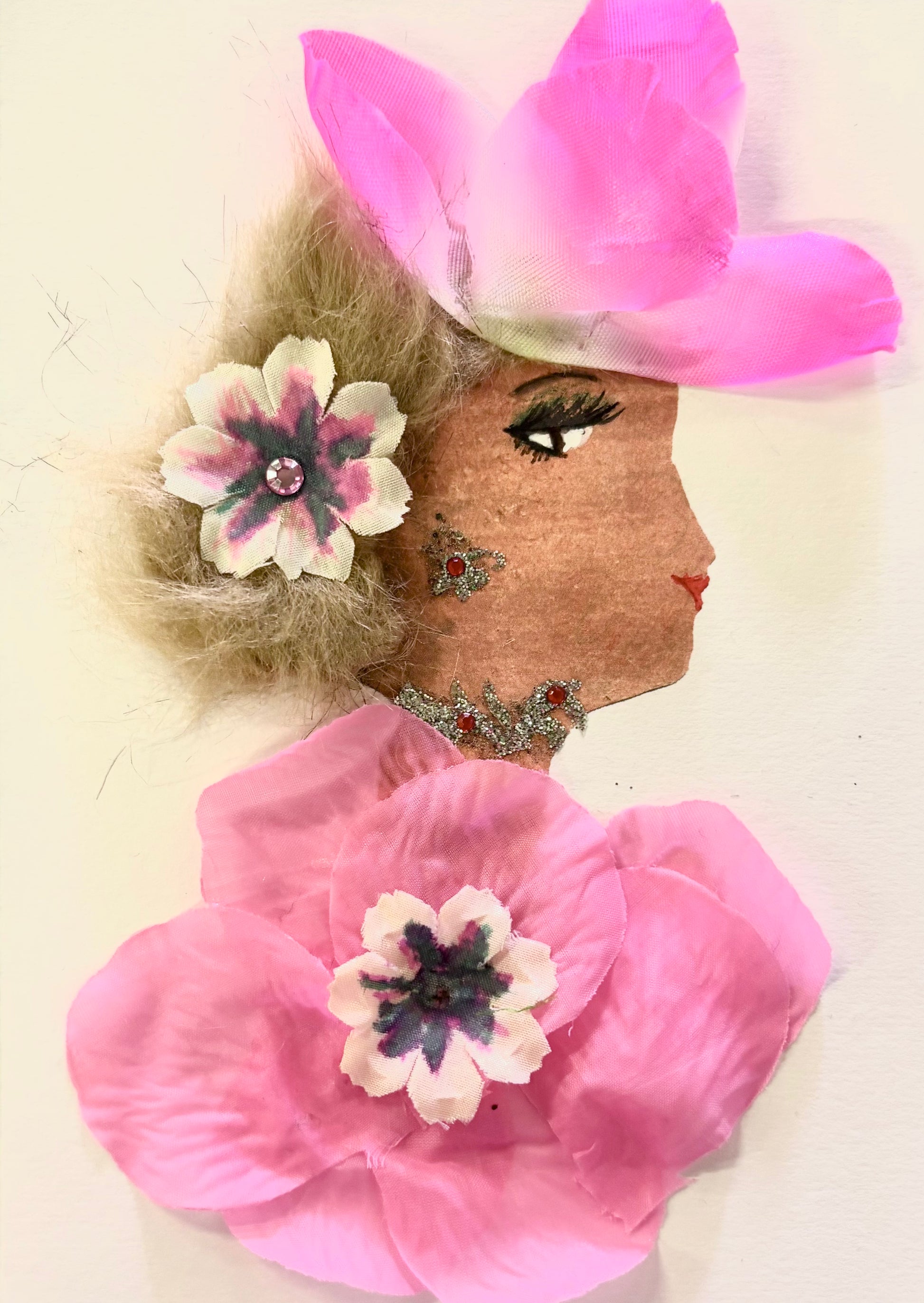 Handmade portrait of a woman in profile. She has light brown curly hair with a pink and white flower in it, and wears a large pink flower hat. Her dress is made from pink fabric flowers, and she has glittery red and silver decorations on her face and neck.