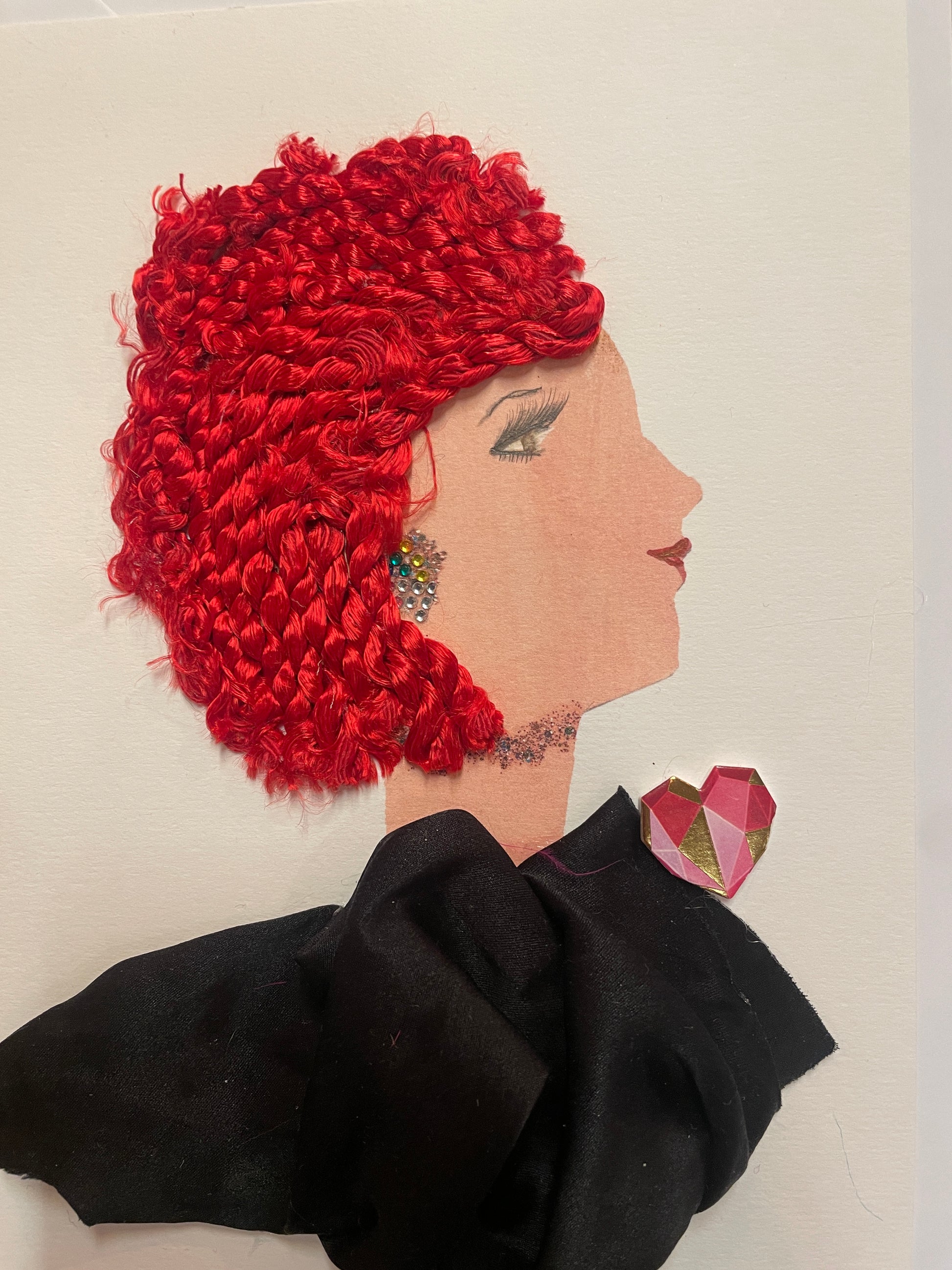 A handmade card of a woman in profile with bright red textured hair, a multi-coloured gem earring, and a black outfit with a pink and gold patterned heart near her shoulder.