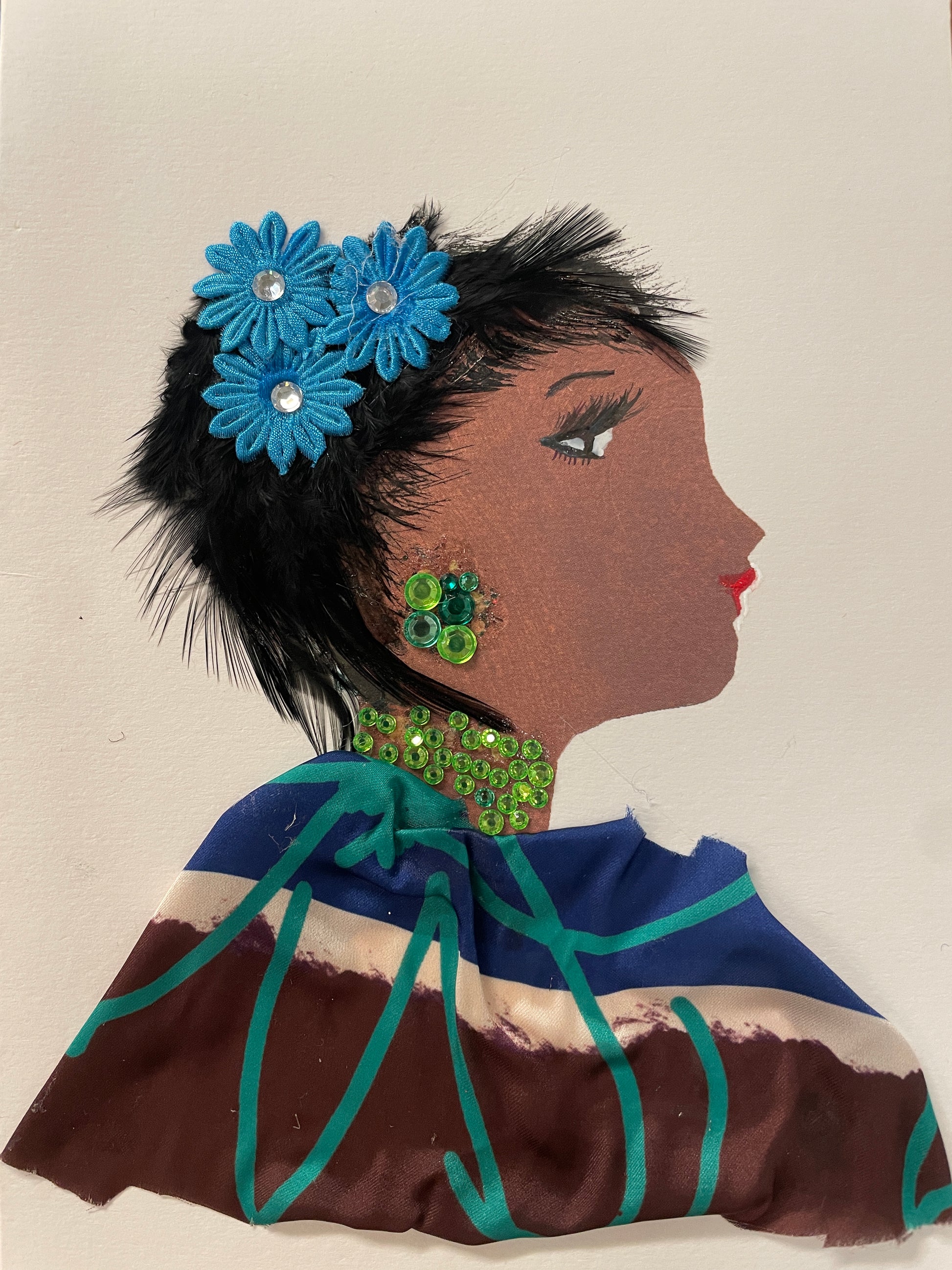 This is a handmade card of a woman named Shakira Gardenia. She wears black feathered hair with blue fabric flowers, green sequin jewellery, and a flowing dress in deep hues.