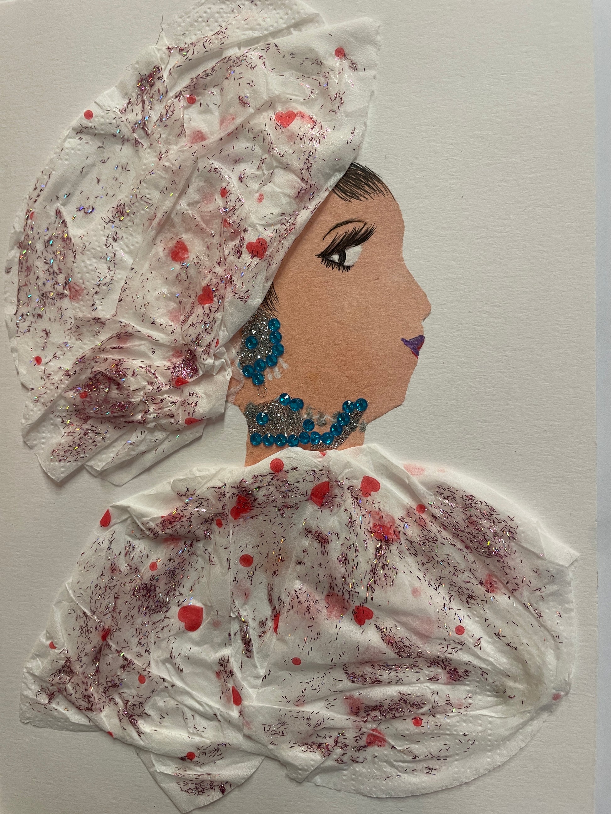 A handmade card showing a woman in profile wearing a white outfit with pink glitter and hearts. She has bright blue earrings and a necklace, and a head wrap made from matching white material.