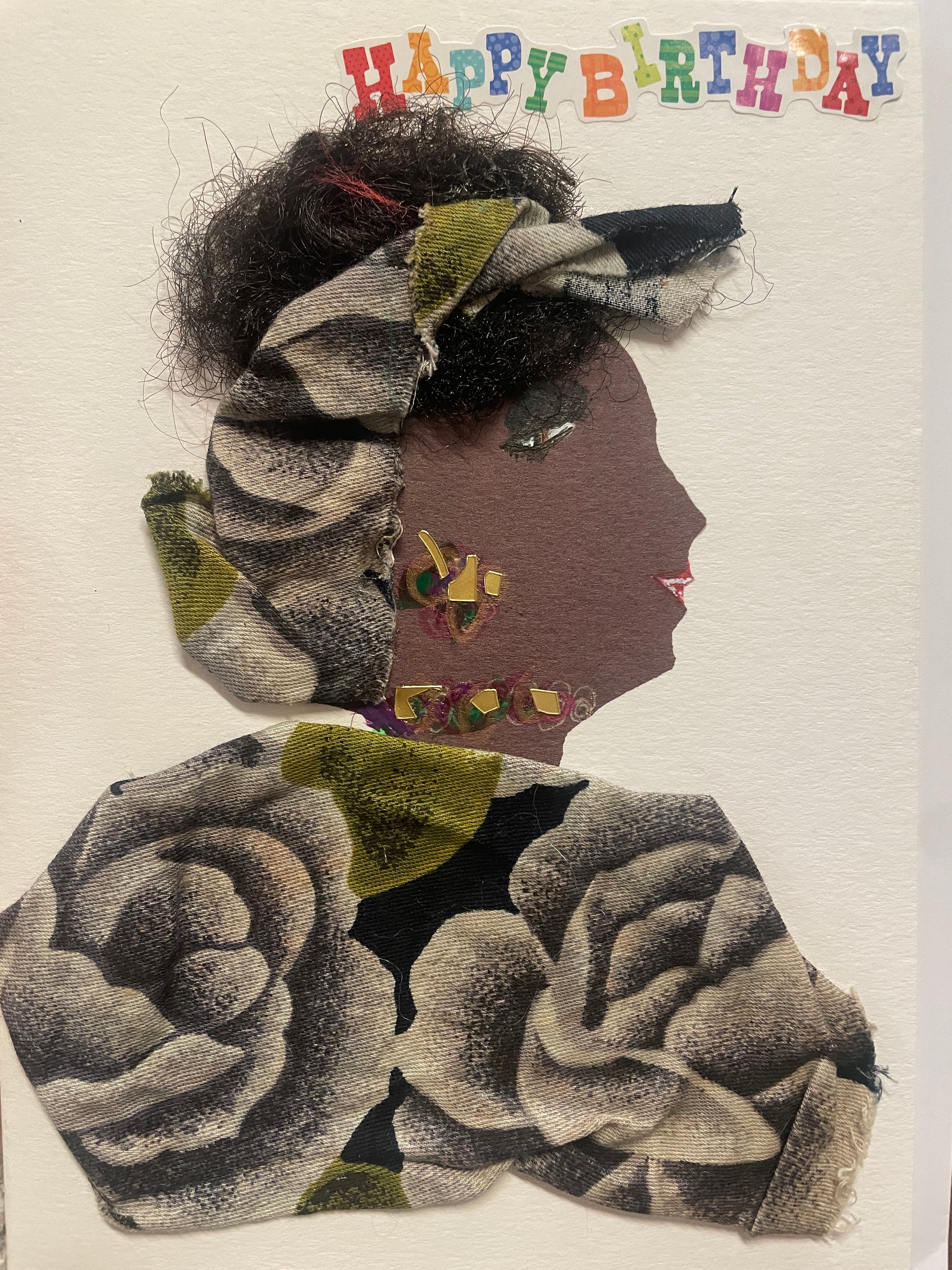 This is a handmade card of a woman named Temi Tope. Her hair is styled with black textured fibres and wrapped in floral fabric that matches her outfit. She wears a necklace and earring made of multicoloured gems. Her outfit is crafted from fabric featuring large grey roses with touches of green. At the top, colourful letters spell out “Happy Birthday.”