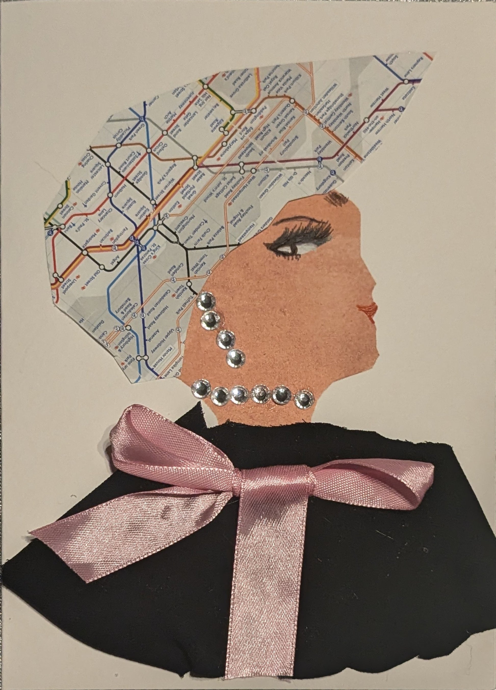 I handmade this card of a woman named Tube Map. She’s shown in profile with dramatic lashes, red lips, and a headwrap made from a section of the London Underground map. Her jewellery is created from silver rhinestones cascading from ear to neck. She wears a sleek black outfit topped with a neatly tied pale pink satin bow.