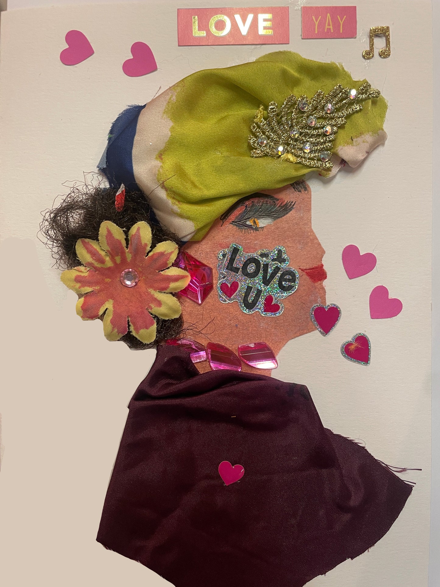 A handmade card of a woman in profile wearing a maroon dress, a yellow-green and navy head wrap with gold decoration, a pink flower on her shoulder, and surrounded by pink heart stickers and the word “LOVE”.