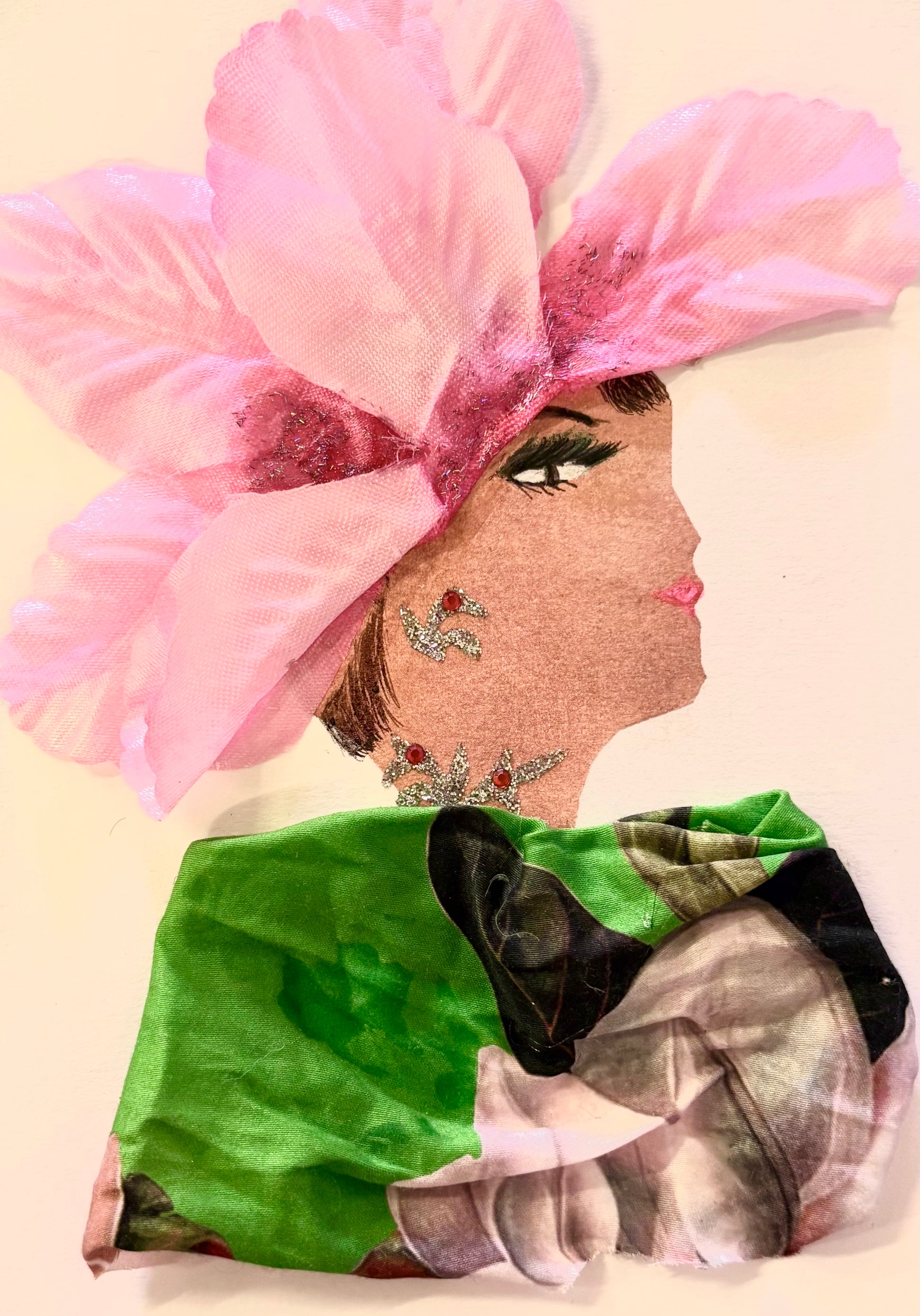 Handmade portrait of a woman in profile. She has a large pink fabric flower on her head, green eye makeup, and pink lipstick. She wears a green and floral-patterned outfit and has silver and pink decorations on her ear and neck.
