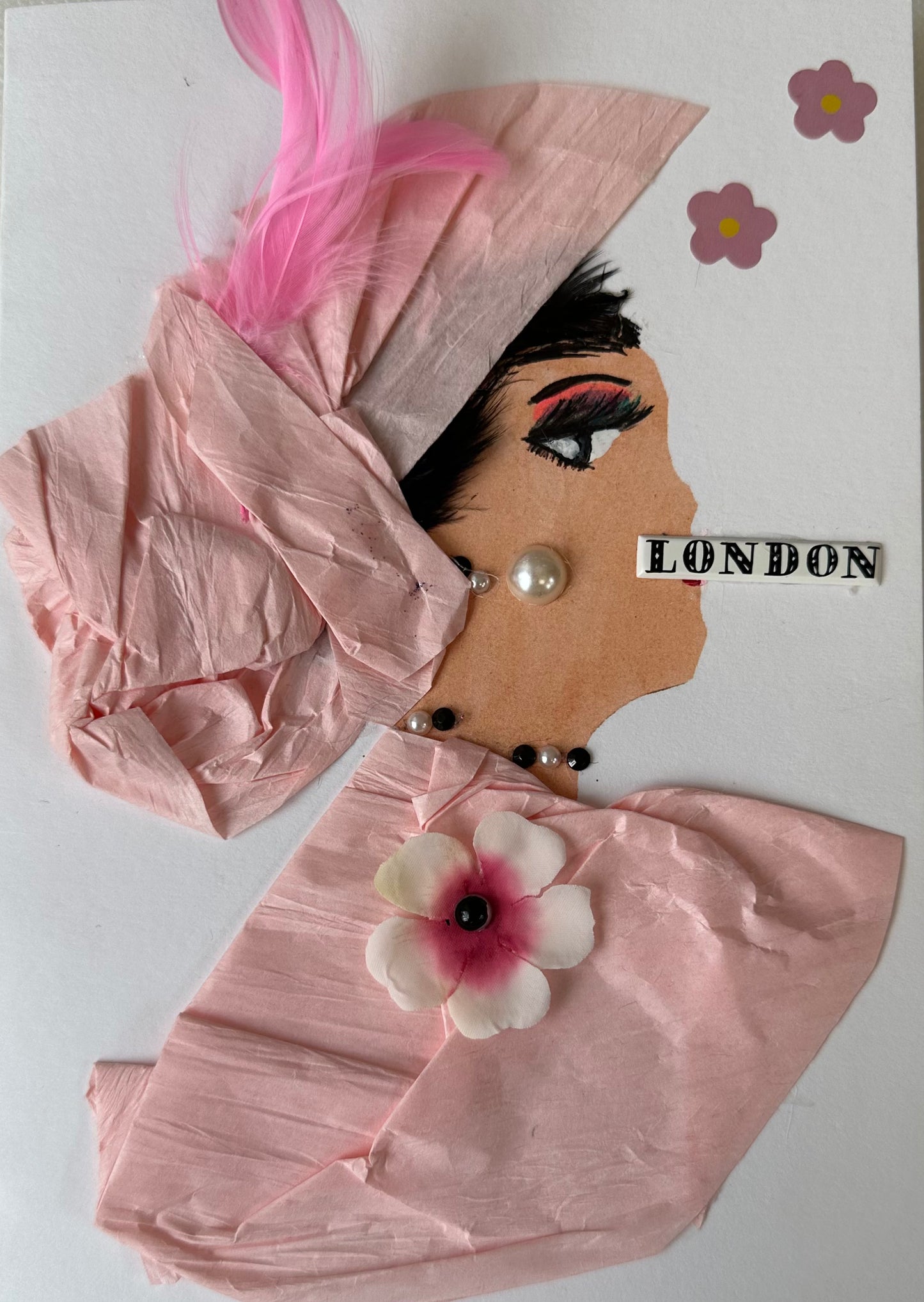 This handmade card that I created is over London Lily. She is dressed in a light pink blouse and hair piece. Her blouse has a cherry blossom attached, and a pink feather in her hair. She has pearl like earrings and London written across her mouth. There are also more flowers on the card.
