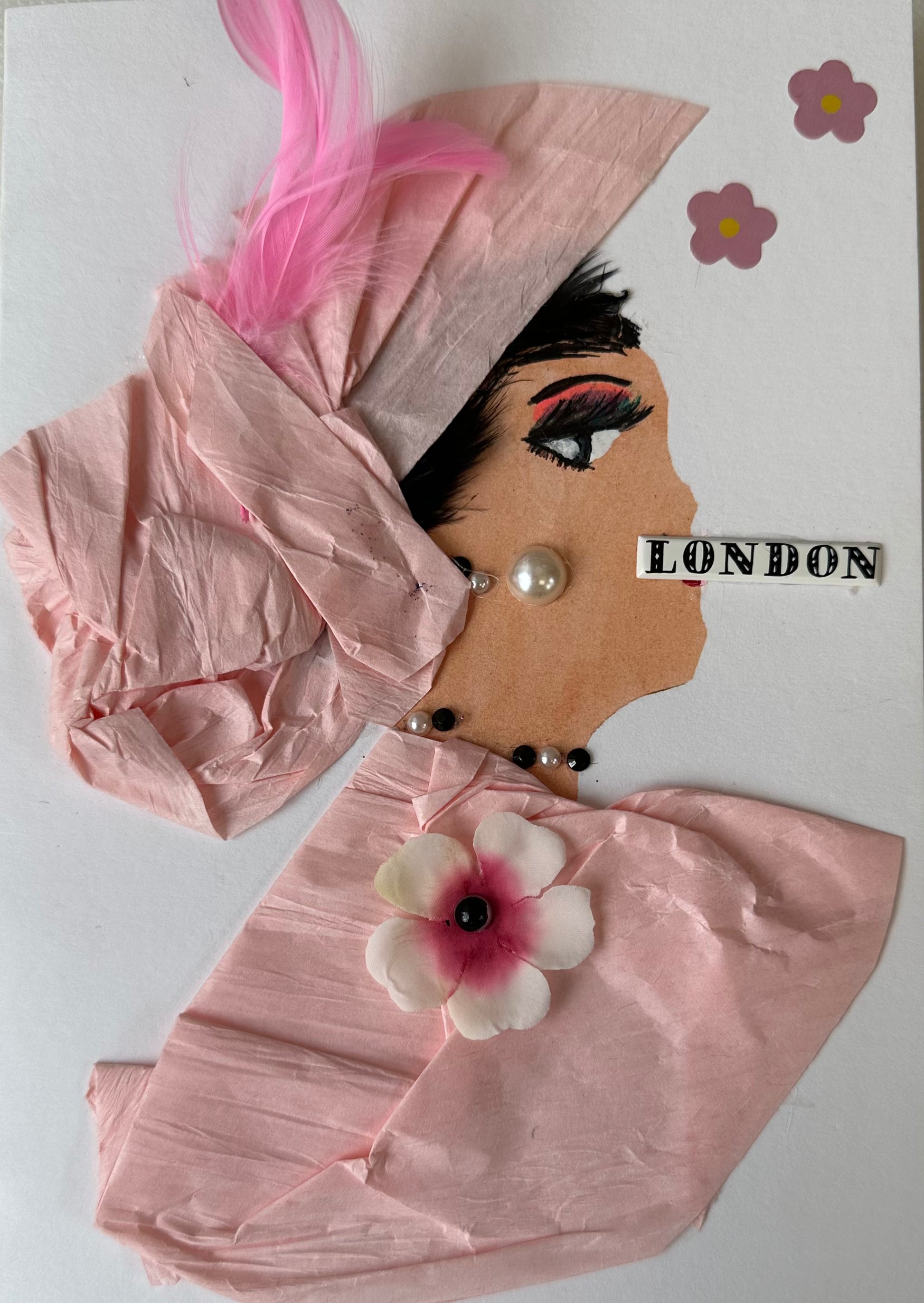 This handmade card that I created is over London Lily. She is dressed in a light pink blouse and hair piece. Her blouse has a cherry blossom attached, and a pink feather in her hair. She has pearl like earrings and London written across her mouth. There are also more flowers on the card.