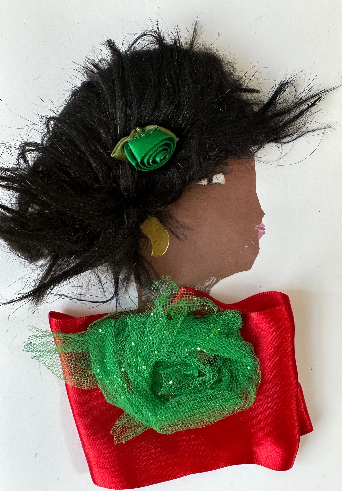 Handmade card showing the side profile of a woman with voluminous black textured hair, adorned with a small green ribbon rose. She wears a gold circular earring and pink lipstick. Her outfit features a red satin fabric with a green glittery netting rosette at the neckline. The background is plain white.