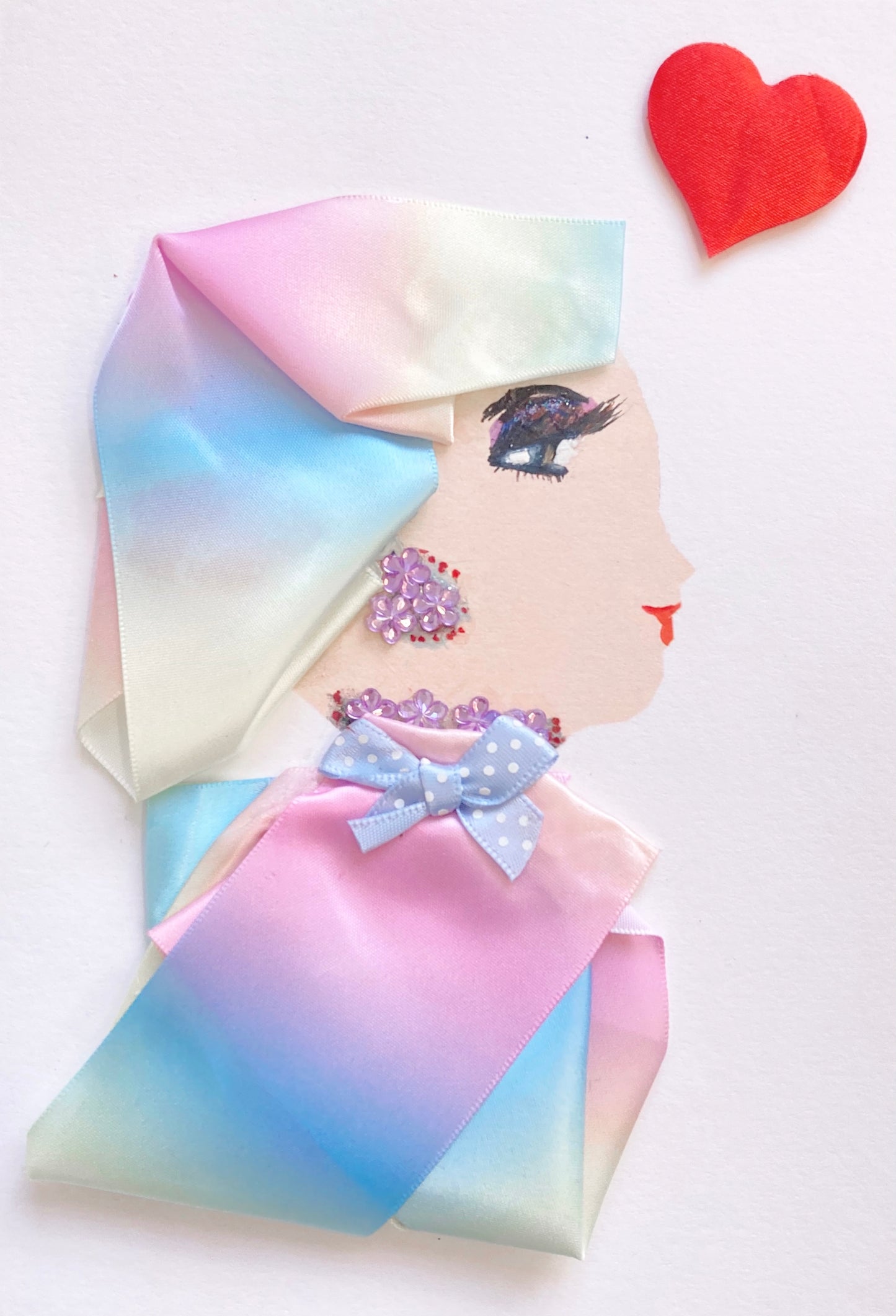 This card is called Dr Nora. She wears a rainbow headdress and blouse, both of which are a silky material. Her jewellery is made of small purple flower gems. In the top right corner, there is a large red heart.