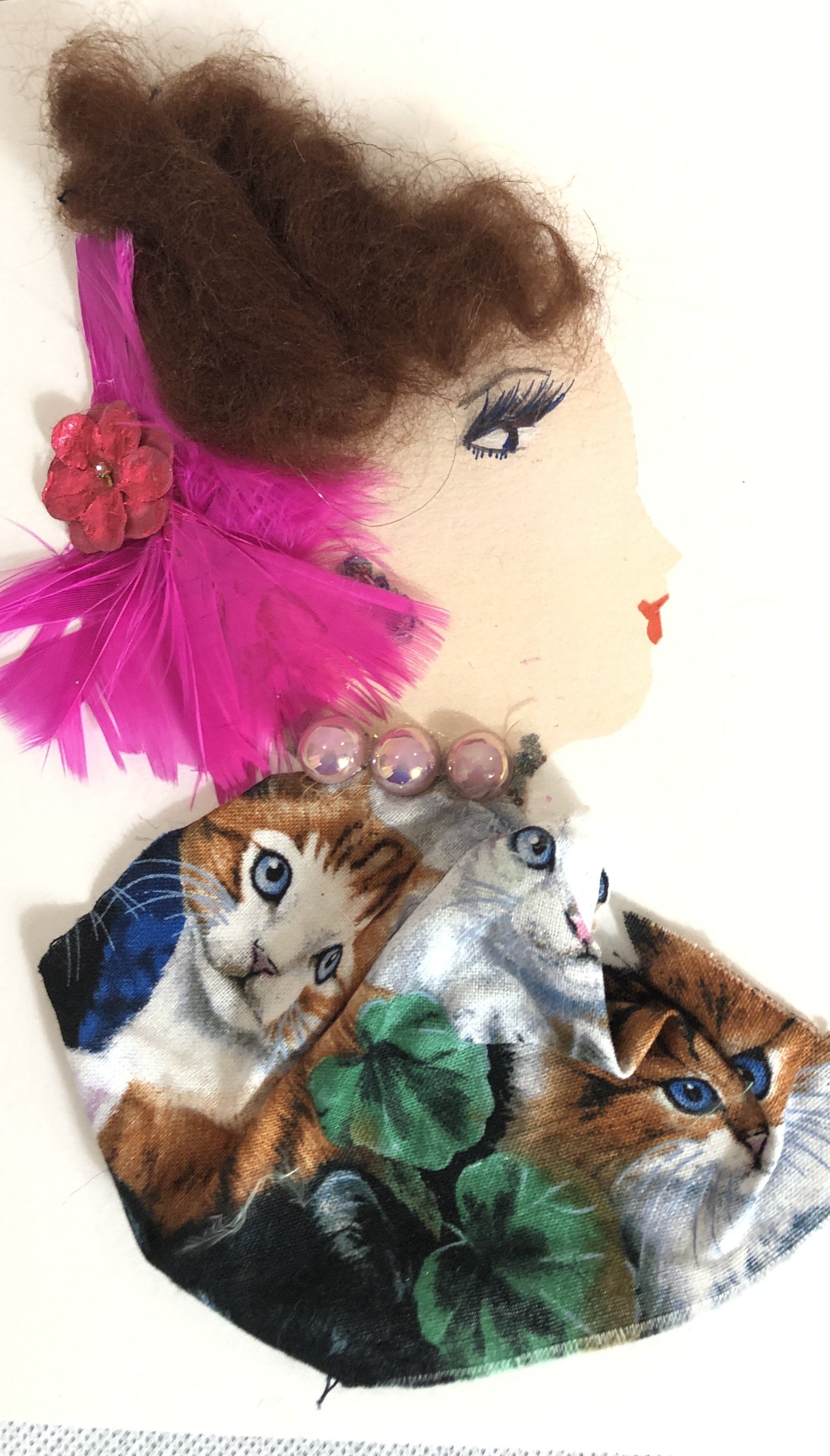 Cathy wears a fabric blouse which has kittens on it. She wears a pearl necklace, and pink feathers in her light brown hair.
