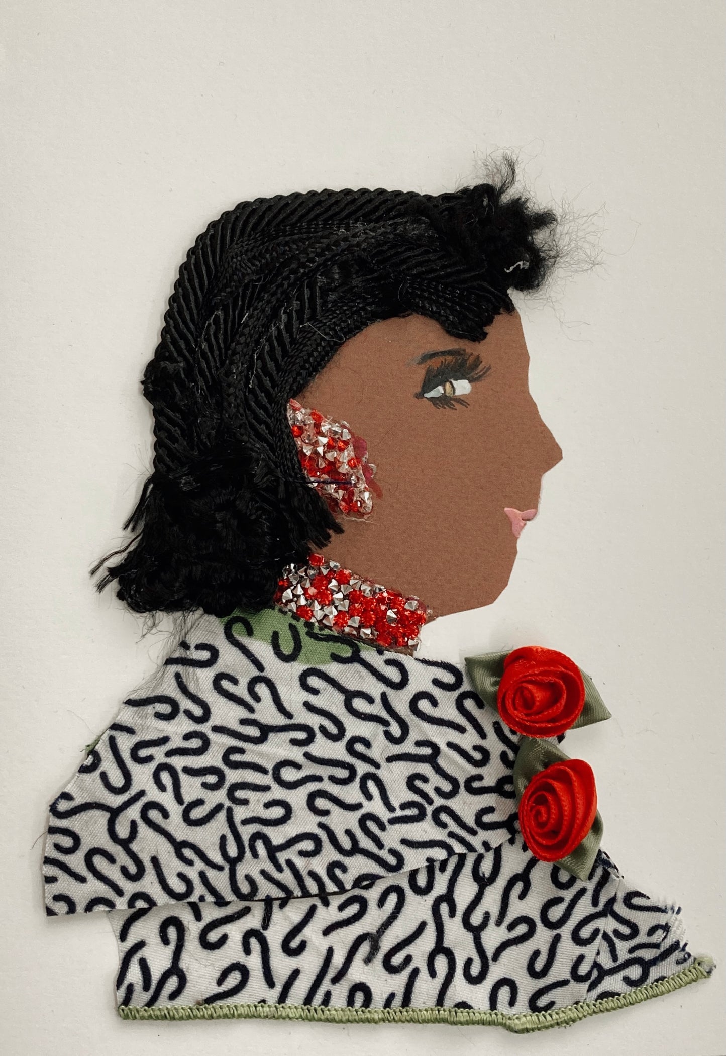 I handmade this card of a woman named Freya Farringdon. She’s shown in profile with detailed black thread hair and soft pink lips. Her jewellery sparkles in red and silver, covering her ear and neckline. She wears a patterned black-and-white fabric outfit, accented with two red satin rosettes and green ribbon leaves.