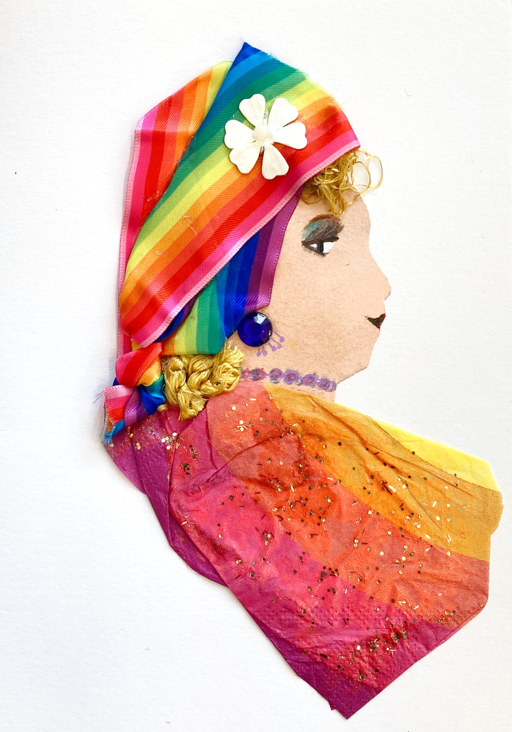 This card is called Kaleidoscopic. She wears a rainbow tissue paper blouse, and a rainbow printed ribbon in her curly blonde hair. She wears a white flower on her headscarf, and a blue earring the same tone as the rainbow.