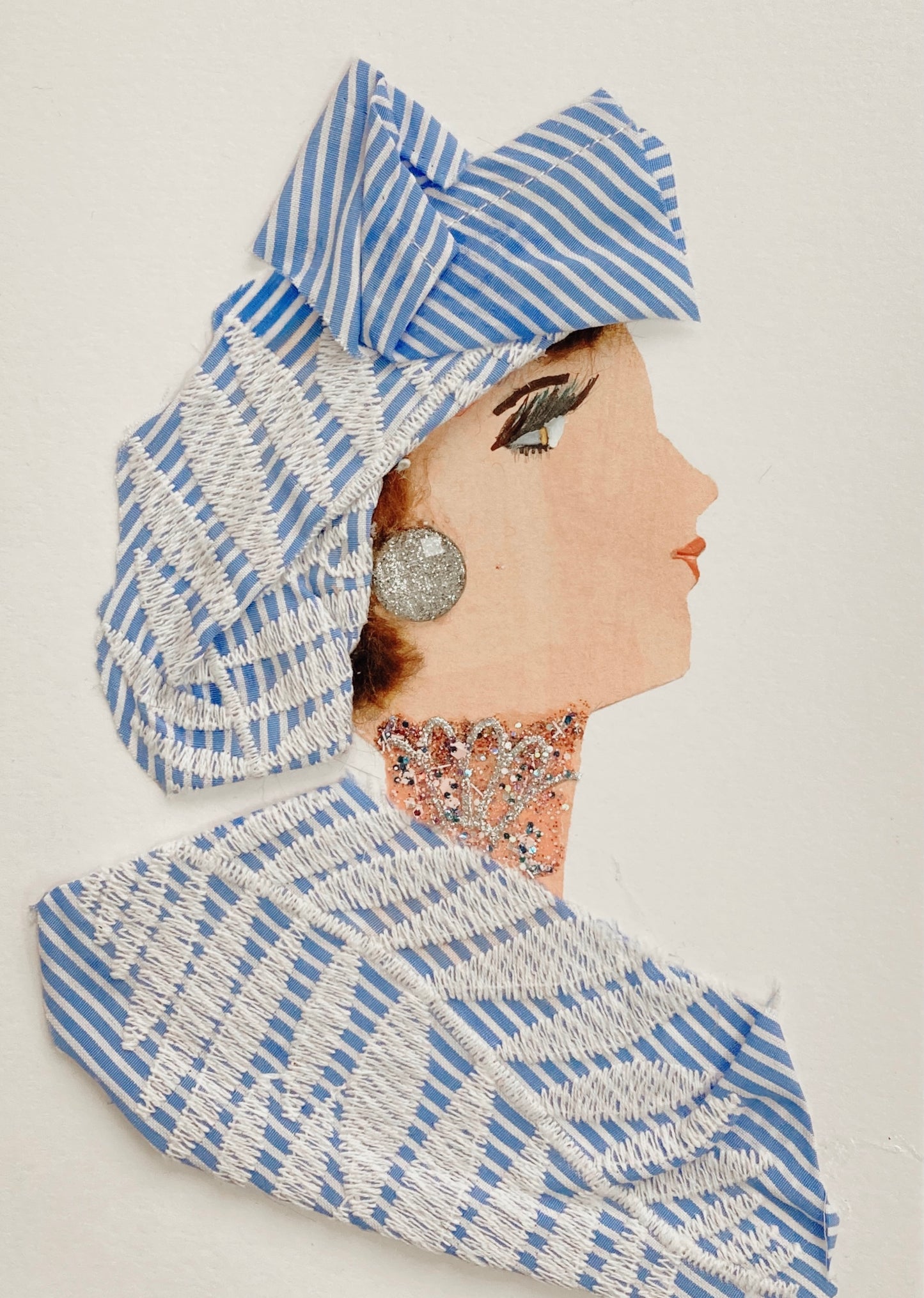 This card has been given the name Robin Redbridge. Robin wears a matching blouse and headscarf, both made of a blue and white pinstripe fabric with a leafy white embroidery overlaying. She wears a large, glittery gem for her earring and a glittery necklace. Visit my website for more