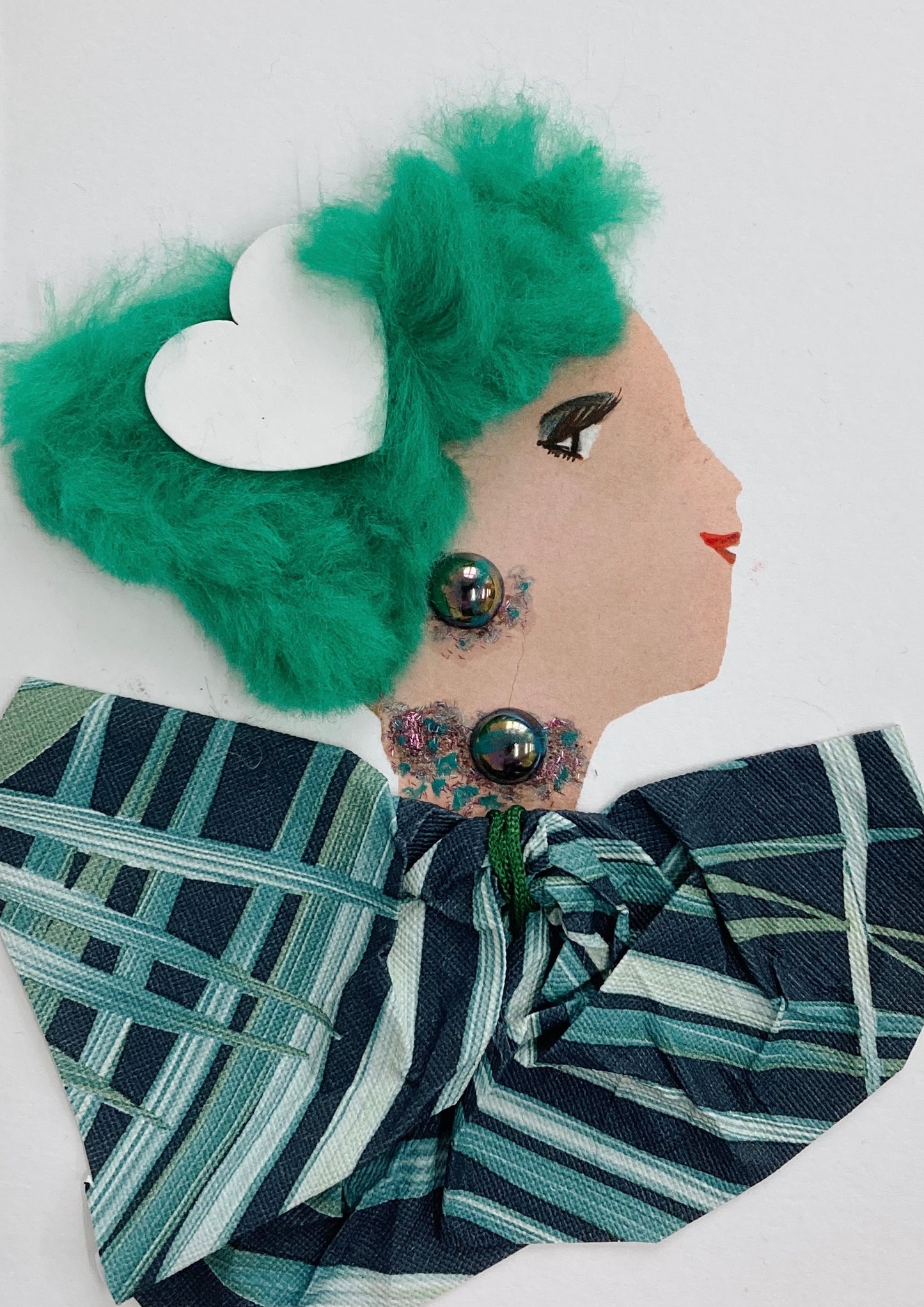 This card has been given the name Nancy. Nancy wears a blouse which has a leafy printed pattern on it. Her hair is furry and green, and has a white heart in it. Her jewellery is an iridescent black pearl earring and necklace.