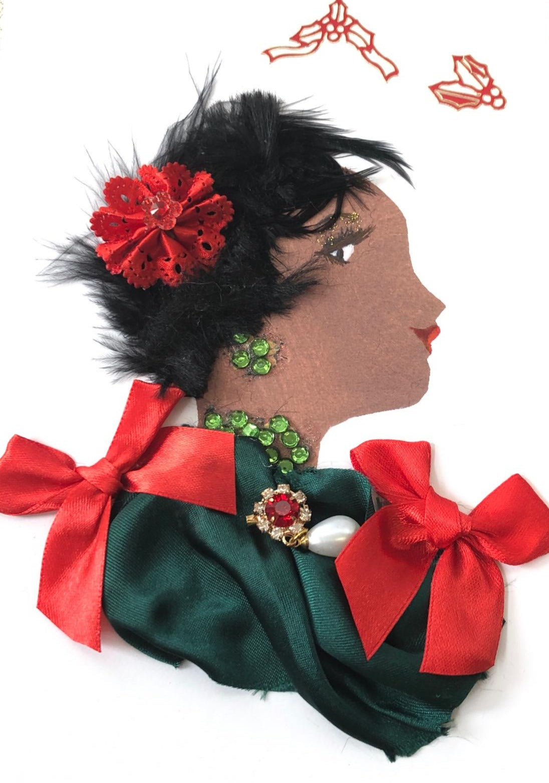 This card has been given the name Temi. Temi wears a forest green silk dress with two red bows. She wears a pendant which has a red crystal at the center and a small pearl hanging from it. In her short feathery black hair, she wears a small red paper flower. In the top right corner, there is two stickers of mistletoe.