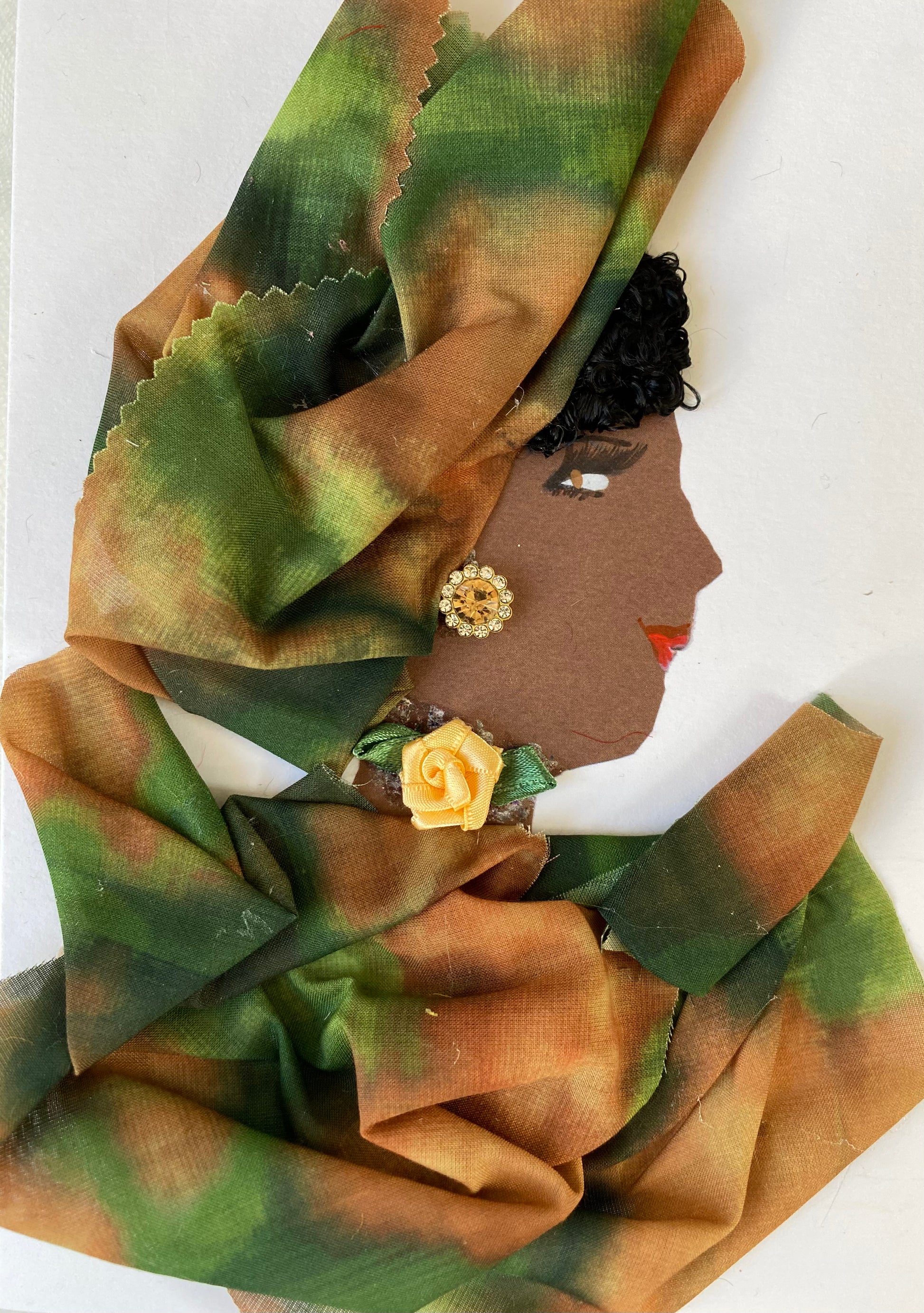 This is a handmade card of a woman named Artistic Annabella. She has dark brown skin and wears a large green and orange wrap that covers her hair and shoulders. Her earring is a shiny golden gem, and there’s a small yellow rose at her neck. Her lips are painted red, and her short black curls peek out from under the wrap.